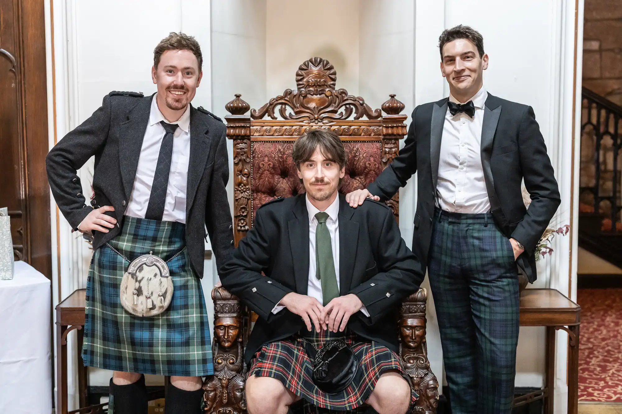 Three men wearing formal attire and kilts, with one seated on an ornate wooden chair and the other two standing on either side.