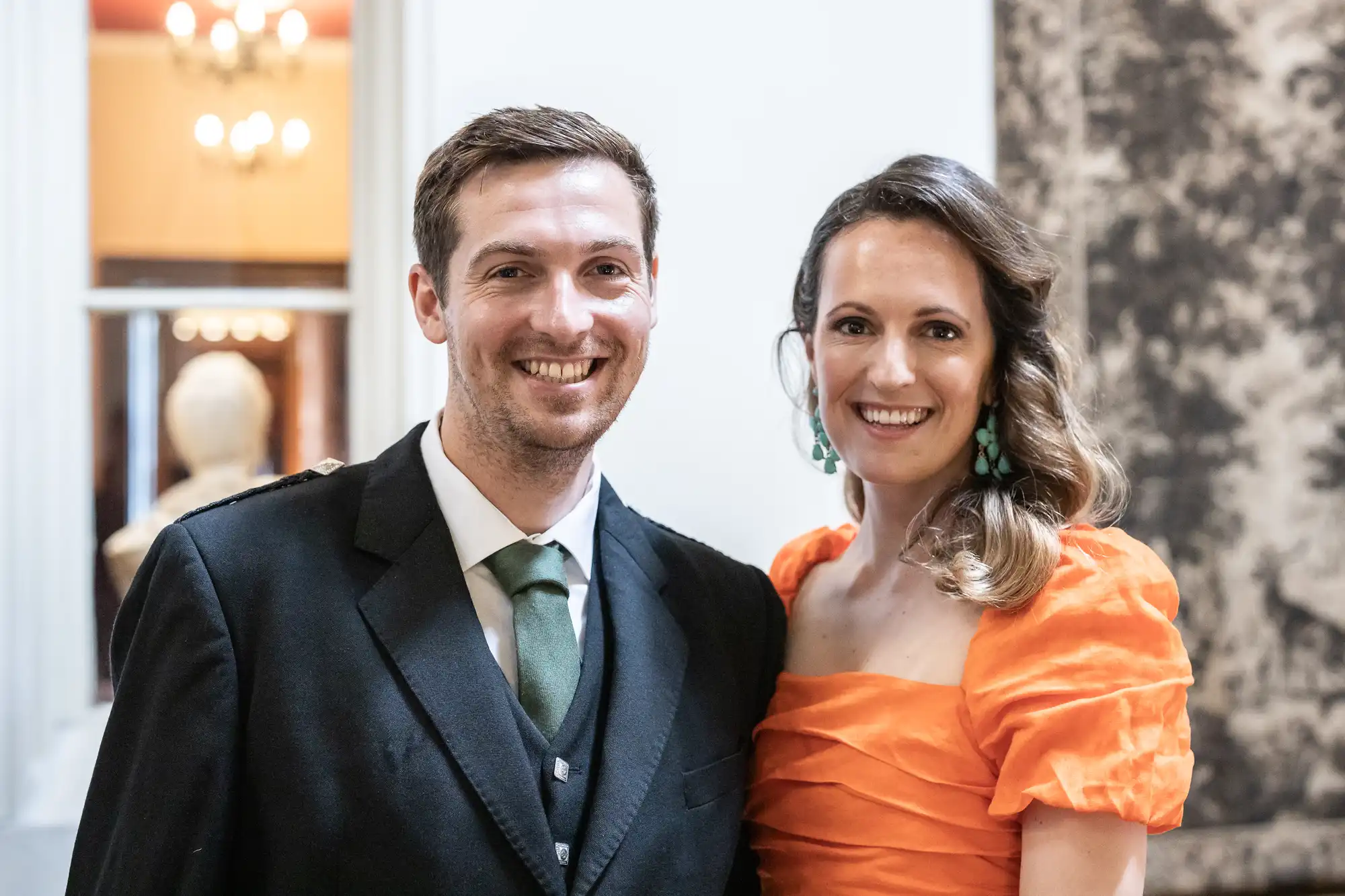 A man and a woman smile at the camera. The man wears a suit and tie, and the woman is dressed in an orange off-the-shoulder top. They are indoors, with blurred lights and decor in the background.