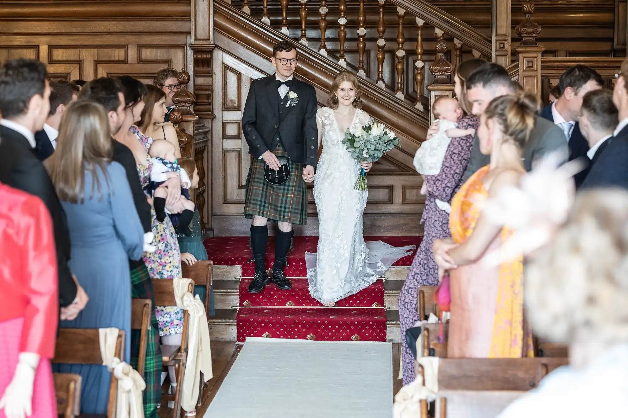 A newlywed couple walks down the aisle together, holding hands, as guests stand on either side in a wooden hall. The groom wears a kilt and the bride holds a bouquet of flowers.