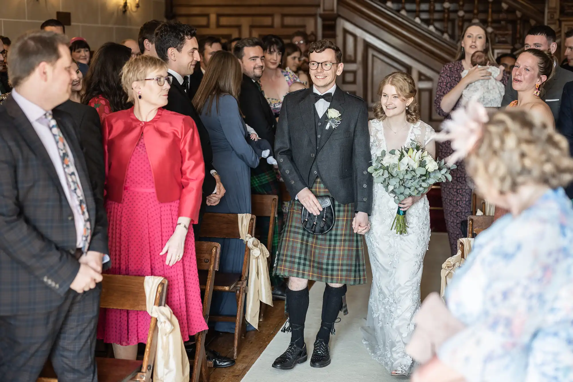 A bride and groom walk down the aisle in a wedding ceremony. The groom is in Scottish attire, and the bride is in a white dress holding a bouquet. Guests are seated and standing nearby, smiling at the couple.