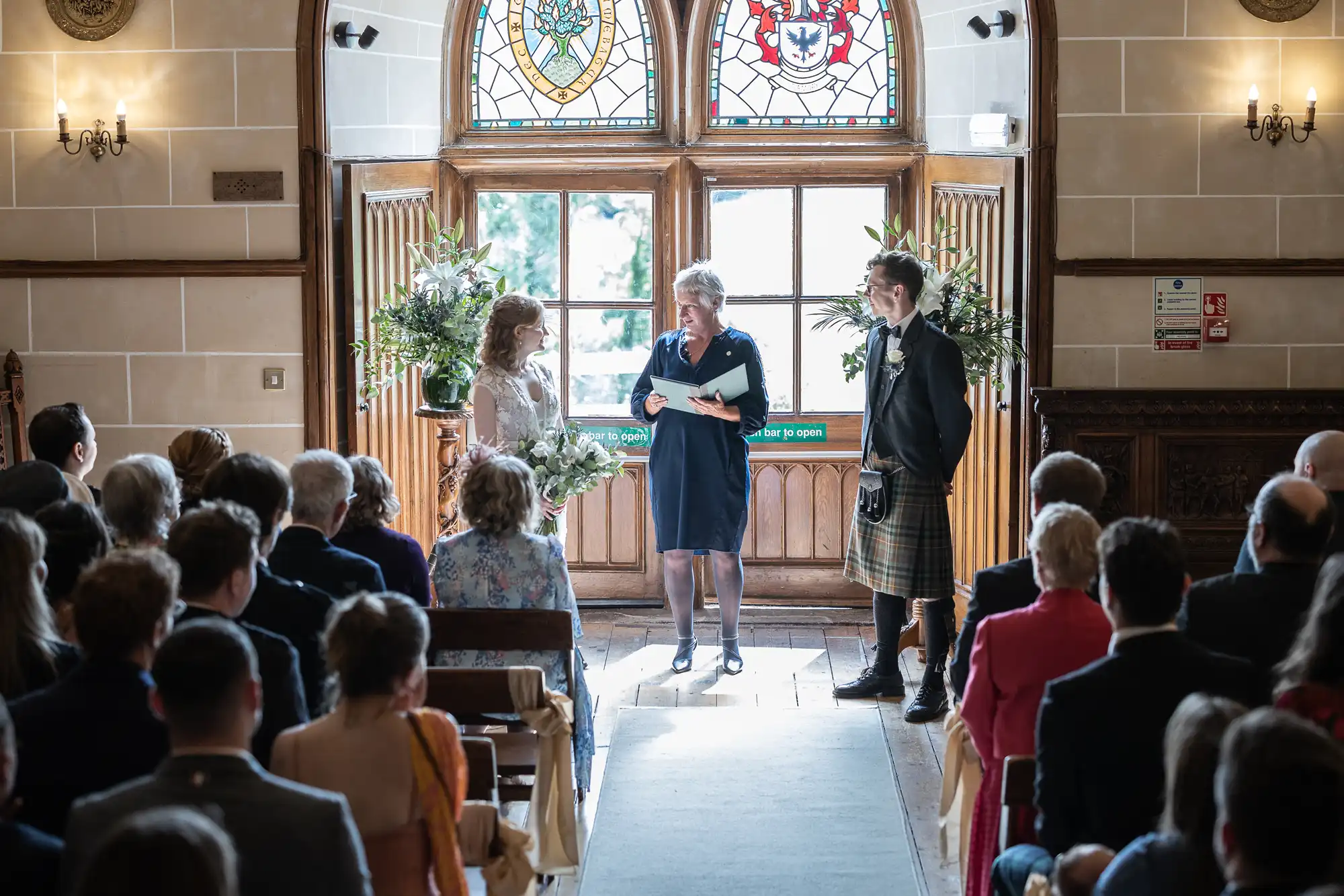 A wedding ceremony takes place in a room with stained glass windows. The officiant stands between the bride holding a bouquet and the groom wearing a kilt, addressing the seated guests.