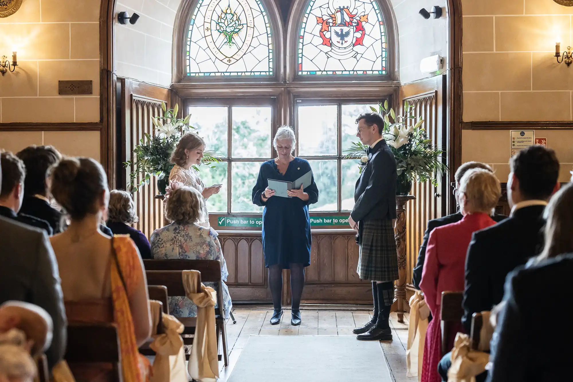 A wedding ceremony taking place indoors with a bride and groom standing at the altar, an officiant reading from a book, and guests seated in chairs decorated with bows.