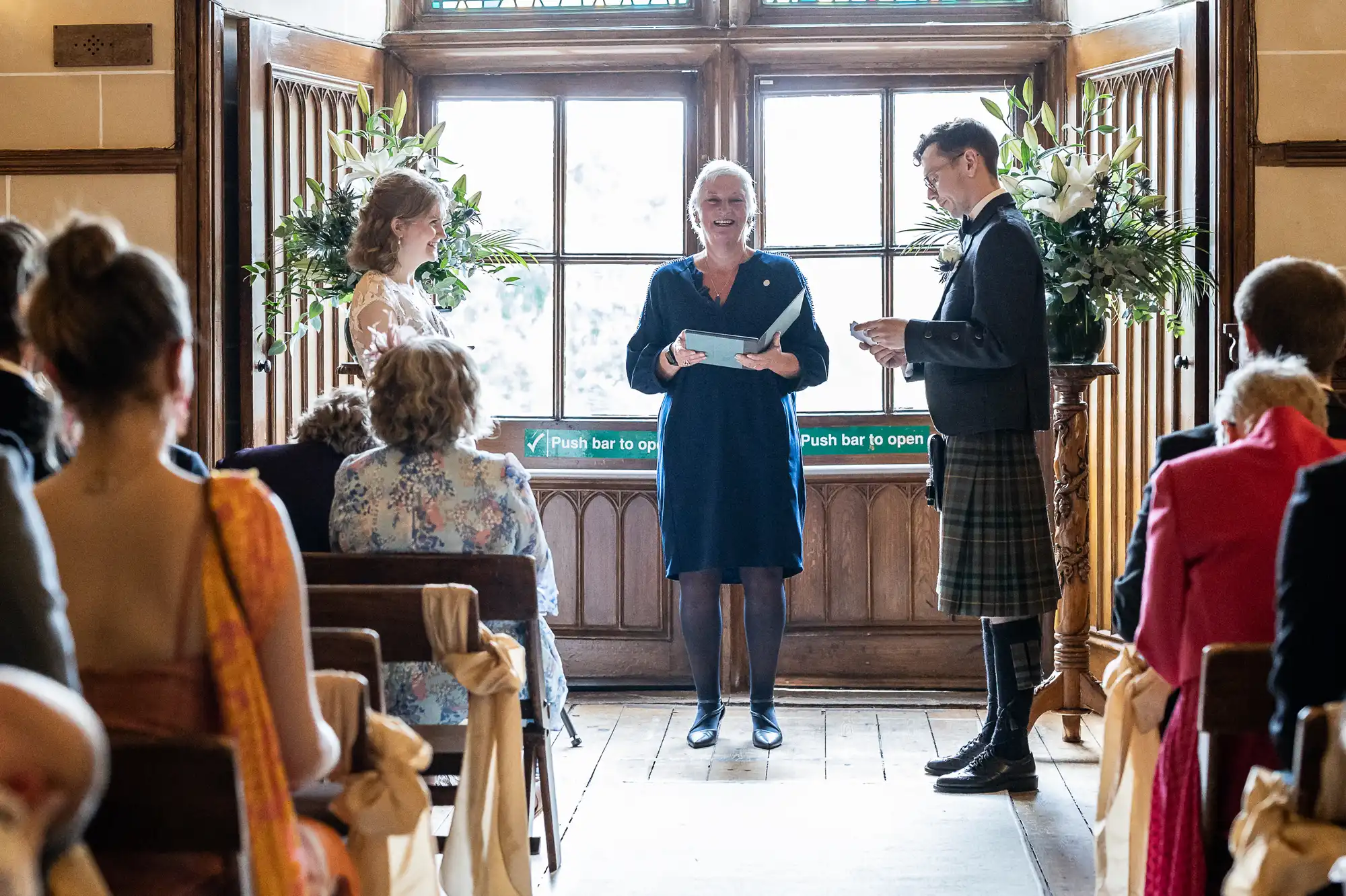 A couple stands facing each other, holding papers, in a well-lit room with large windows. An officiant stands between them, smiling, while guests are seated observing the ceremony.