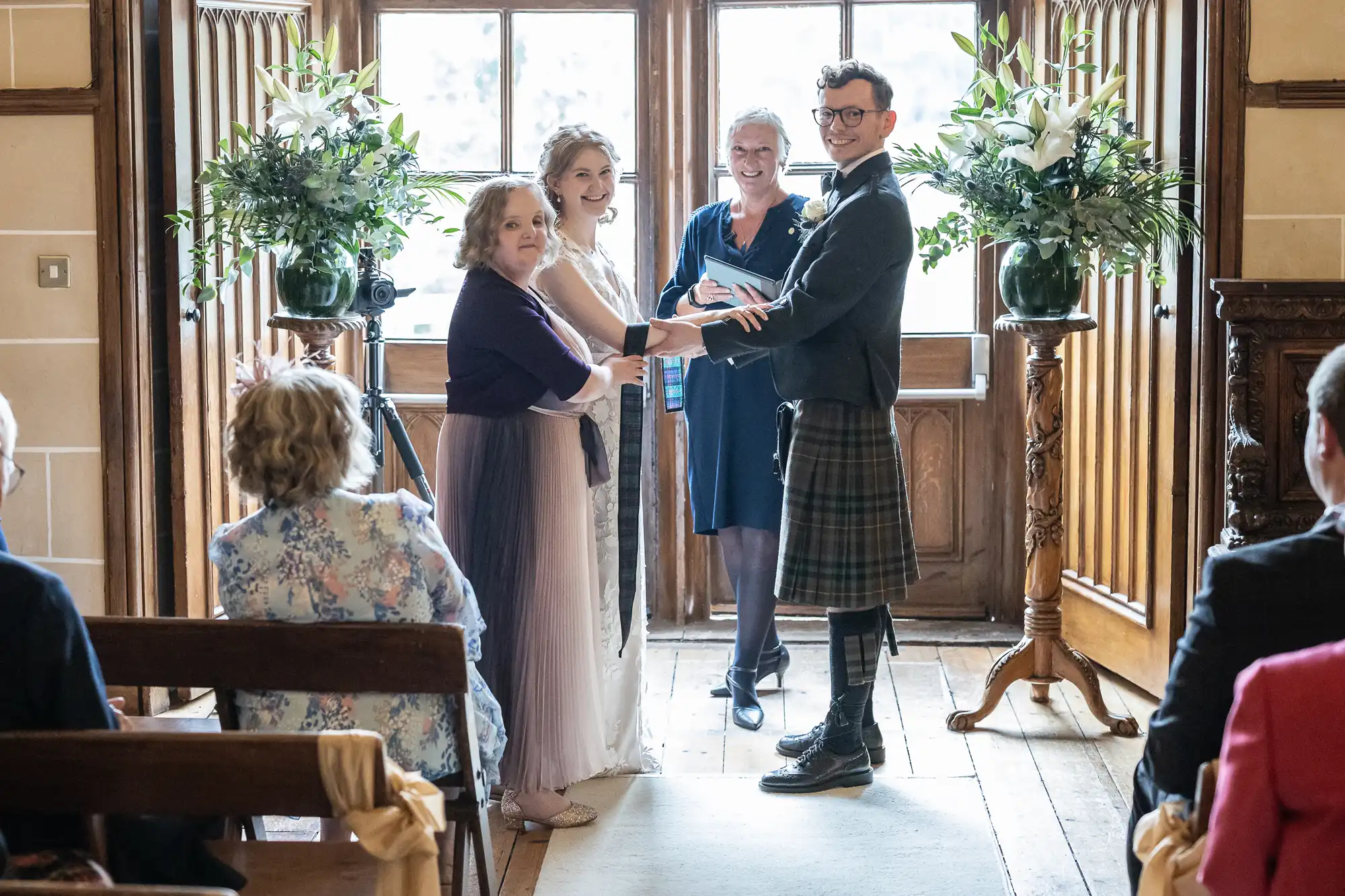 A couple stands at the altar holding hands with an officiant and a woman by their sides. Guests are seated, witnessing the ceremony in a well-lit room with wooden paneling.