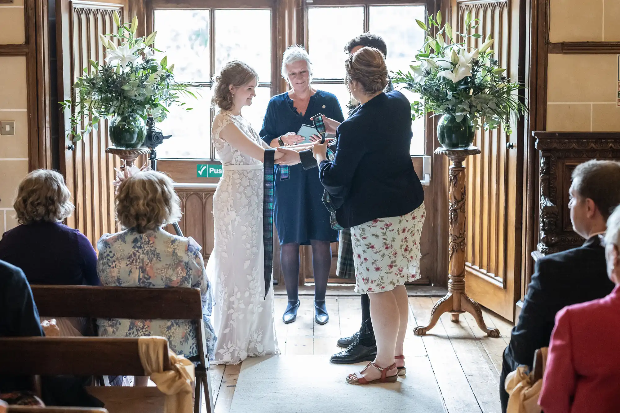 A couple stands at the altar exchanging rings in front of a celebrant and a small audience during a wedding ceremony inside a wood-paneled room with large windows and floral arrangements.