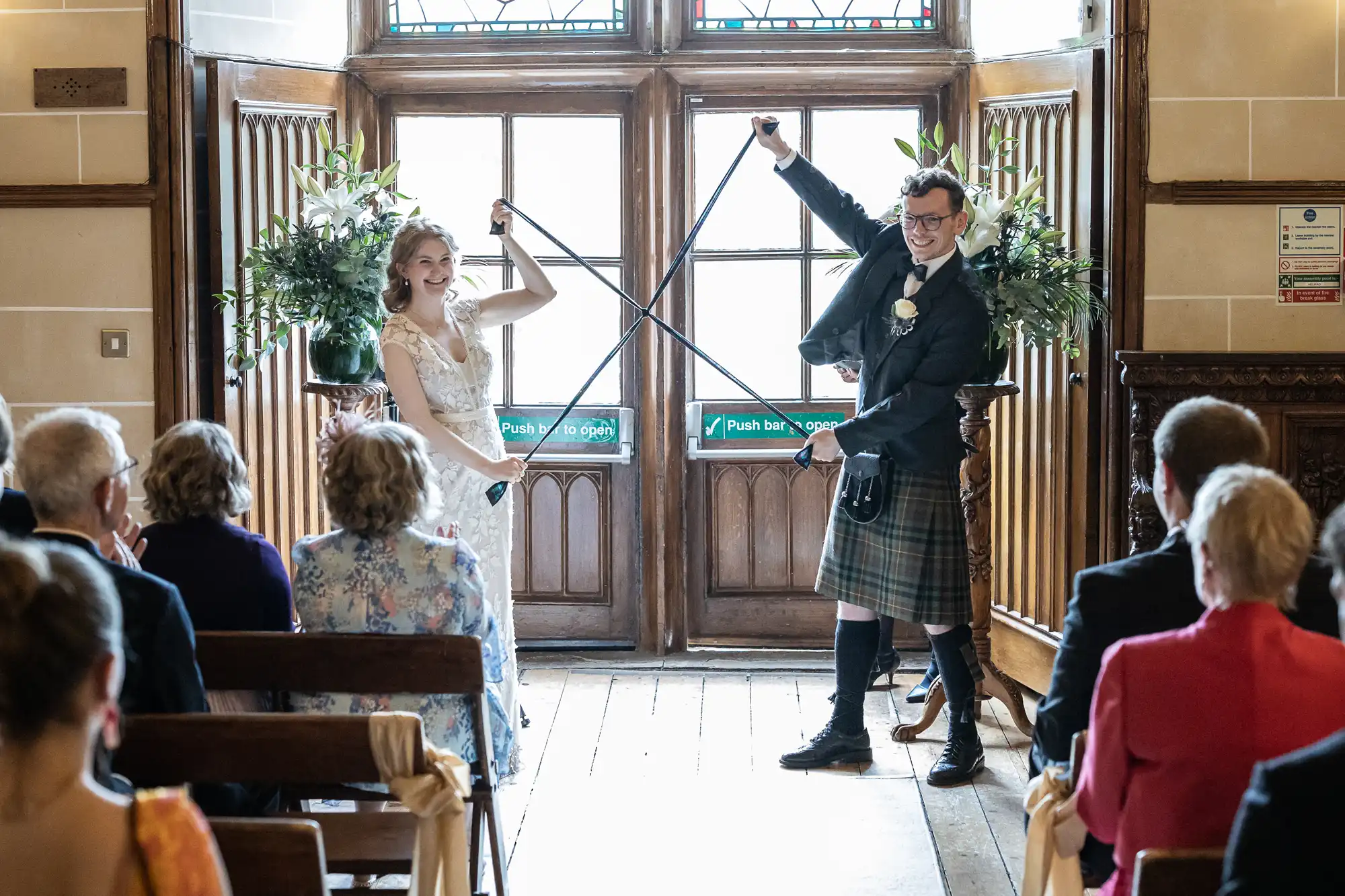 Bride and groom cross swords and smile during their wedding ceremony, standing in front of a wooden door with stained glass and surrounded by seated guests.