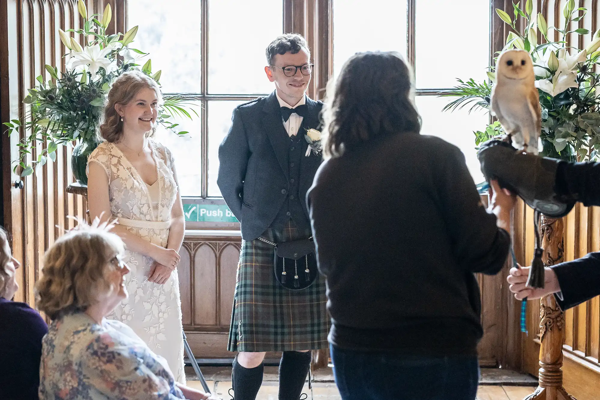 A bride and groom smile as a person holds a perched owl at a wedding ceremony. The groom wears a kilt and the bride is in a white dress. Floral arrangements are visible in the background.