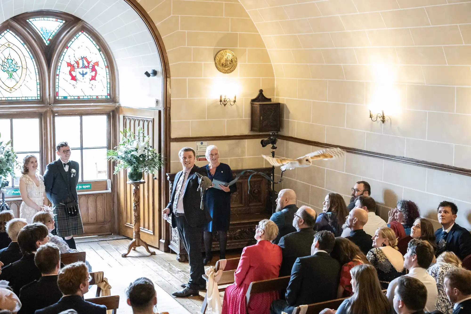 A man is giving a speech in a small room with arched windows, while guests seated in rows listen. A bird is flying near the speaker. A bride and a groom, in a kilt, are standing to the side.