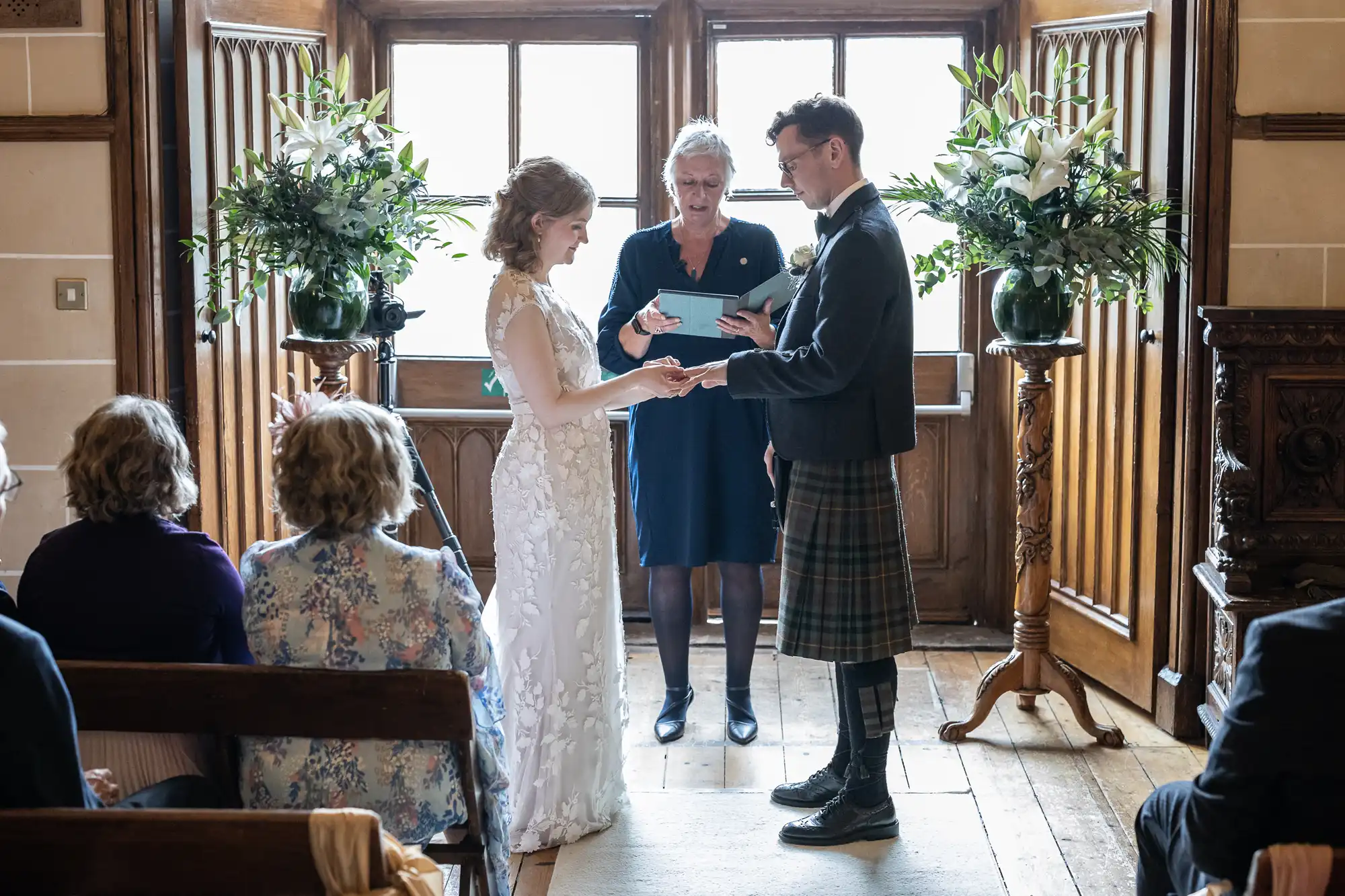 A couple stands facing each other exchanging rings in a wedding ceremony overseen by an officiant. Guests are seated on either side, and large floral arrangements are placed near the couple.