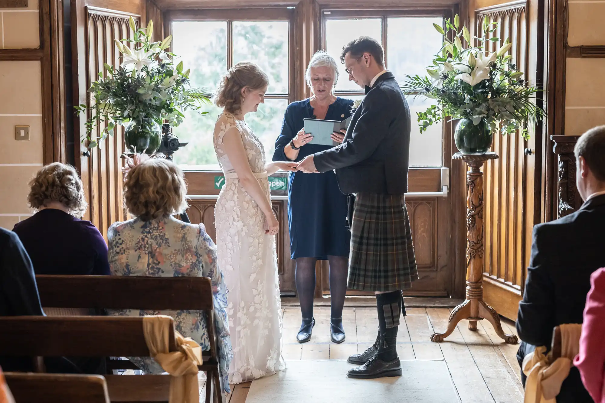 A couple stands before an officiant during their wedding ceremony in a wood-paneled room with large windows and floral arrangements. The bride wears a white dress, and the groom is in a kilt. Guests are seated.