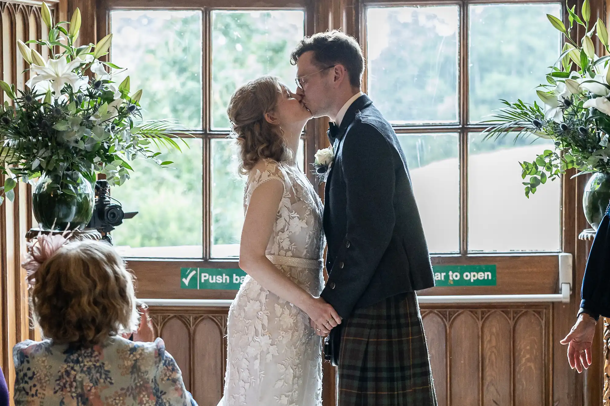 A couple dressed in wedding attire stands kissing in front of a window, surrounded by greenery and floral arrangements, with a visible "Push bar to open" sign on the door behind them.