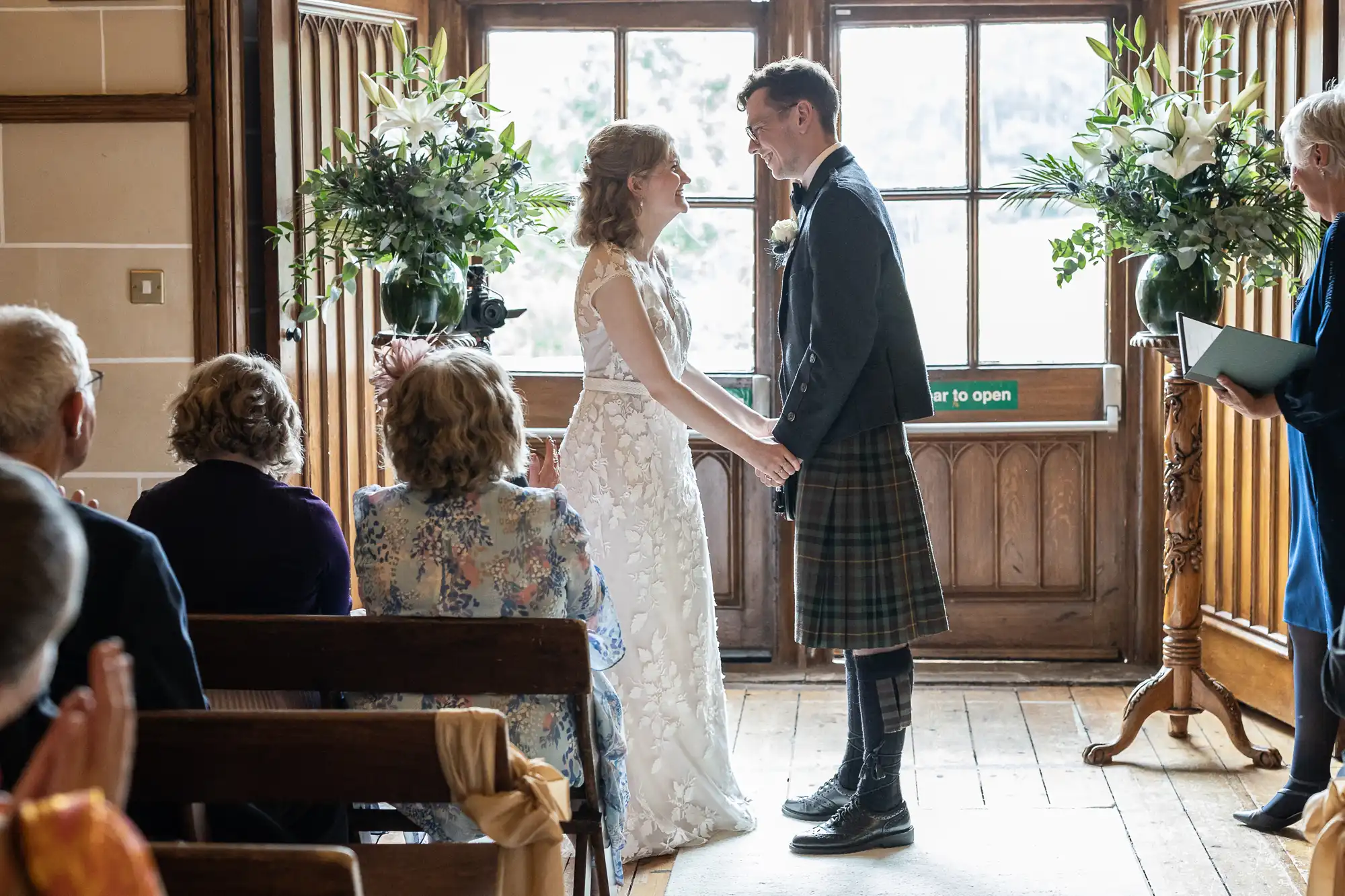 A couple stands at the altar during a wedding ceremony, holding hands and smiling at each other. Guests are seated and watching. Large windows and floral arrangements are in the background.