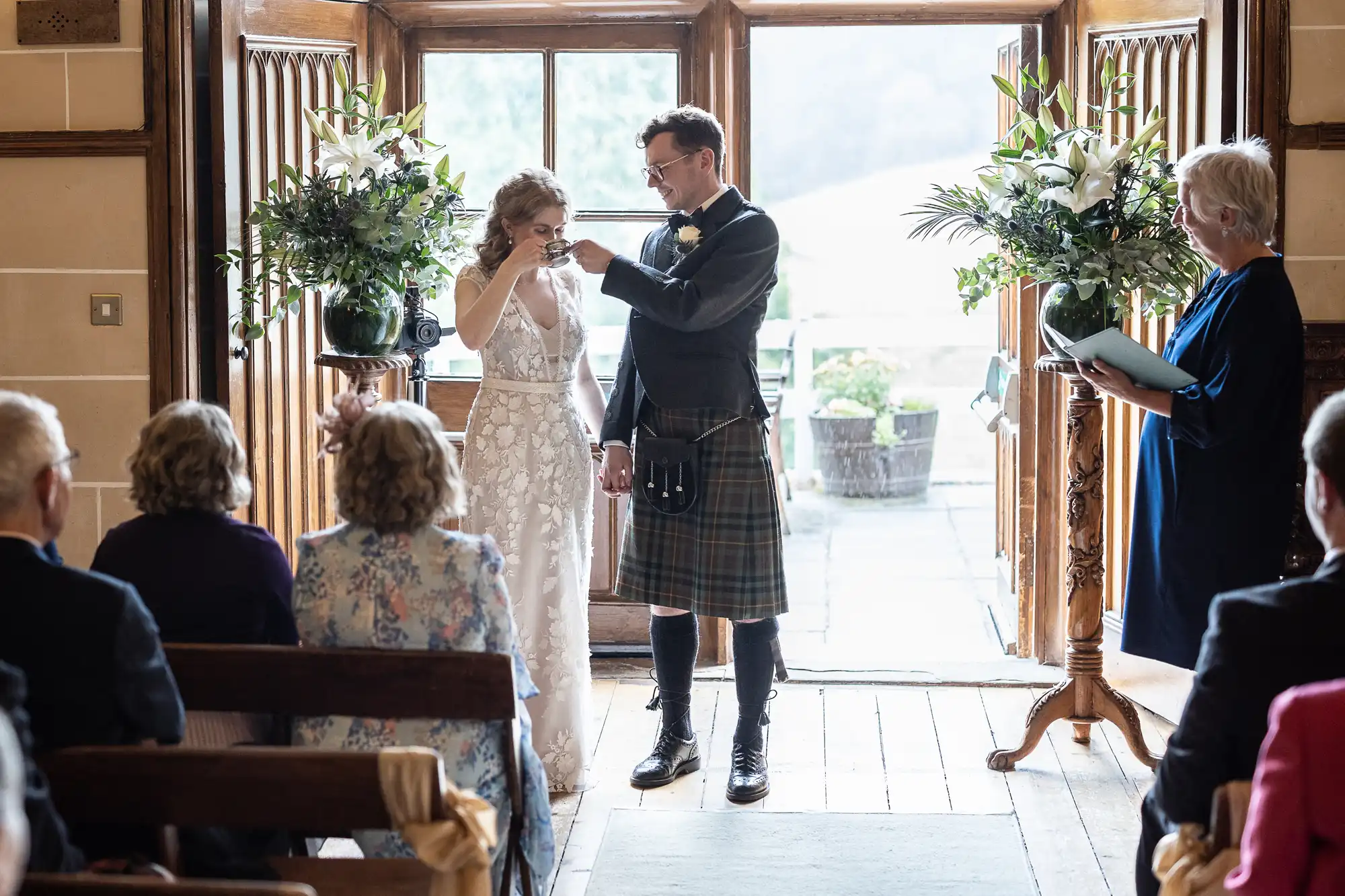 A bride and groom exchange drinks in a ceremony before an audience. The groom is in a kilt, and the bride wears a white dress. A woman stands to the side, reading from a book.