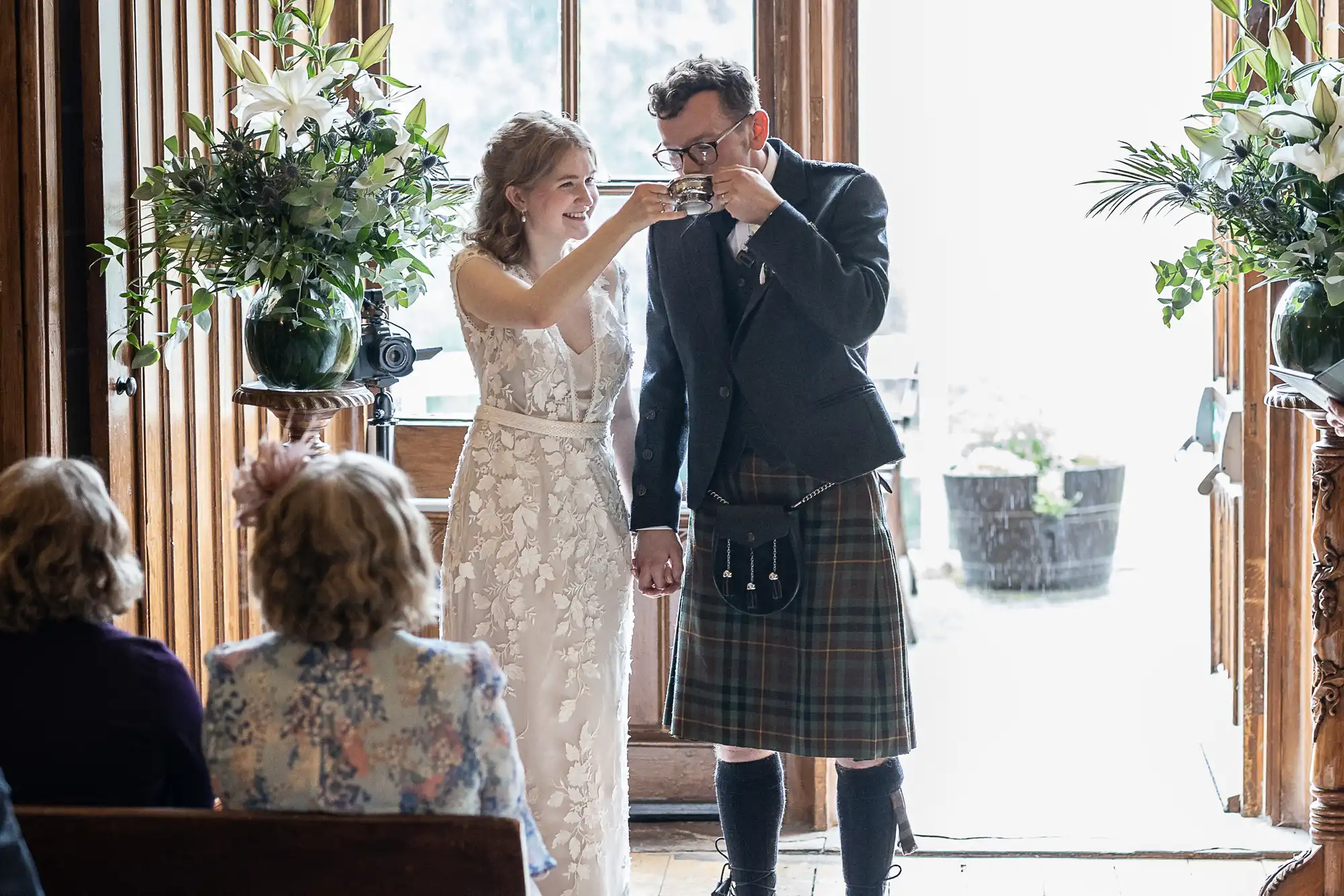 A bride and groom in wedding attire share a drink during their ceremony, with guests seated in the foreground and floral arrangements decorating the scene.