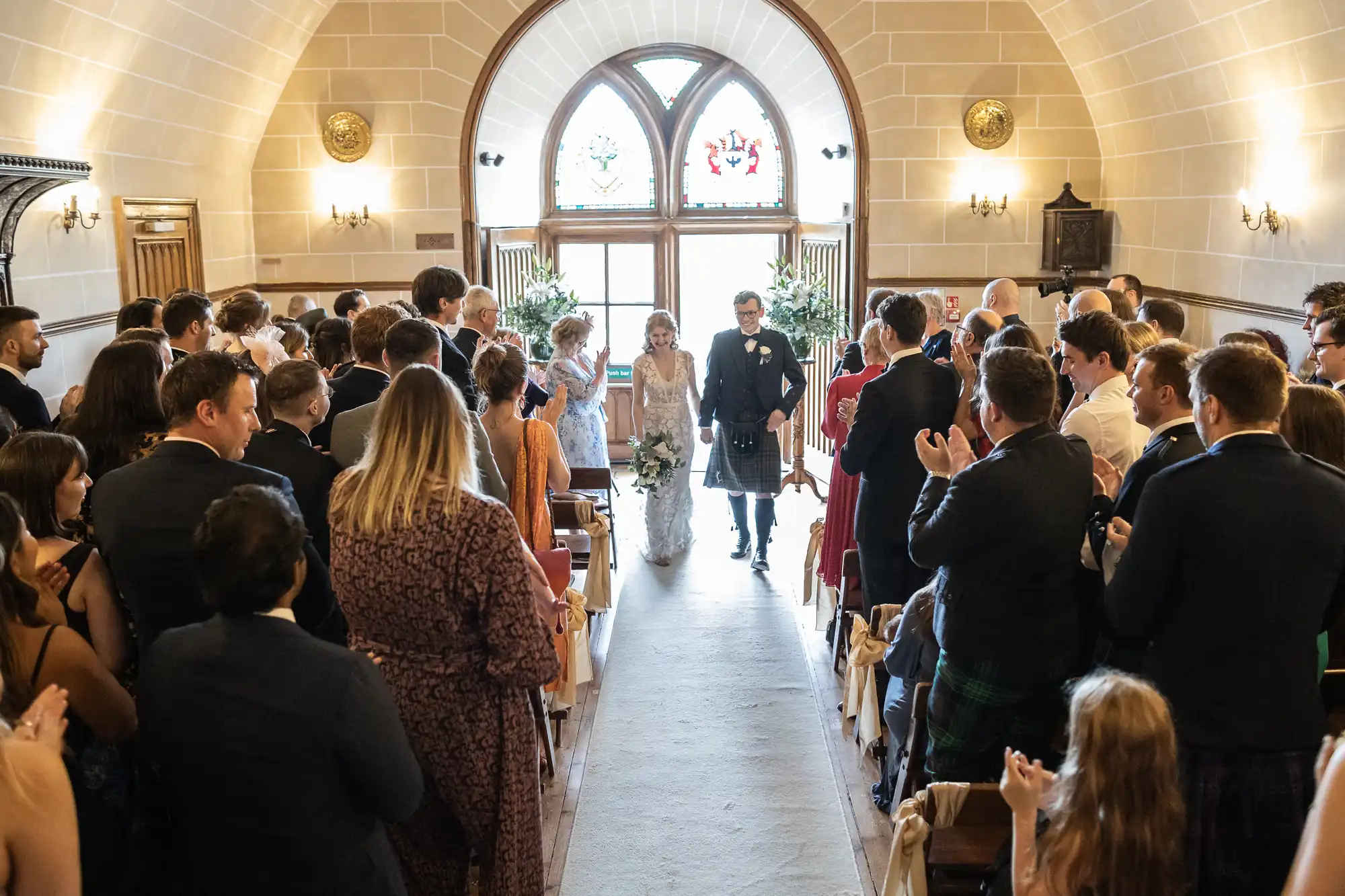 A bride and groom walk down the aisle in a small chapel as guests stand and applaud. The chapel has arched windows and beige stone walls.