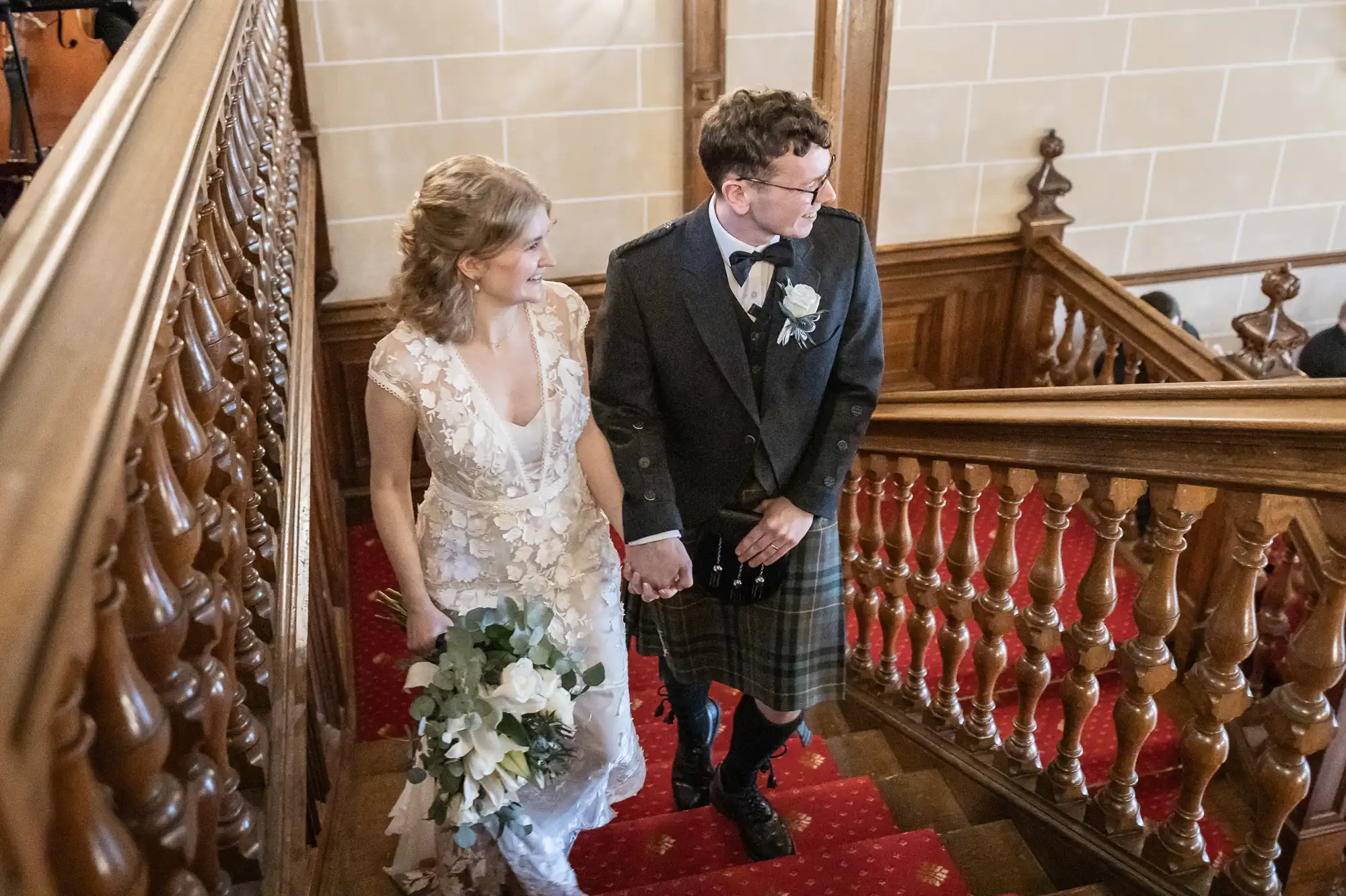 A couple, dressed in wedding attire, holds hands while ascending a wooden staircase. The bride wears a white lace dress and holds a bouquet, while the groom wears a kilt and a jacket.