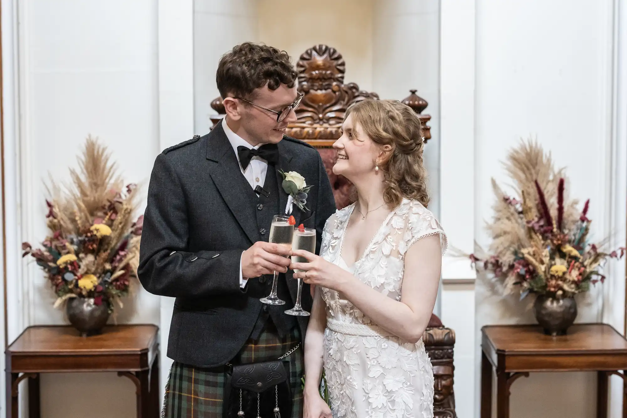 A couple dressed in formal attire toasts with champagne glasses in front of a wooden throne-style chair, flanked by decorative vases with dried floral arrangements.