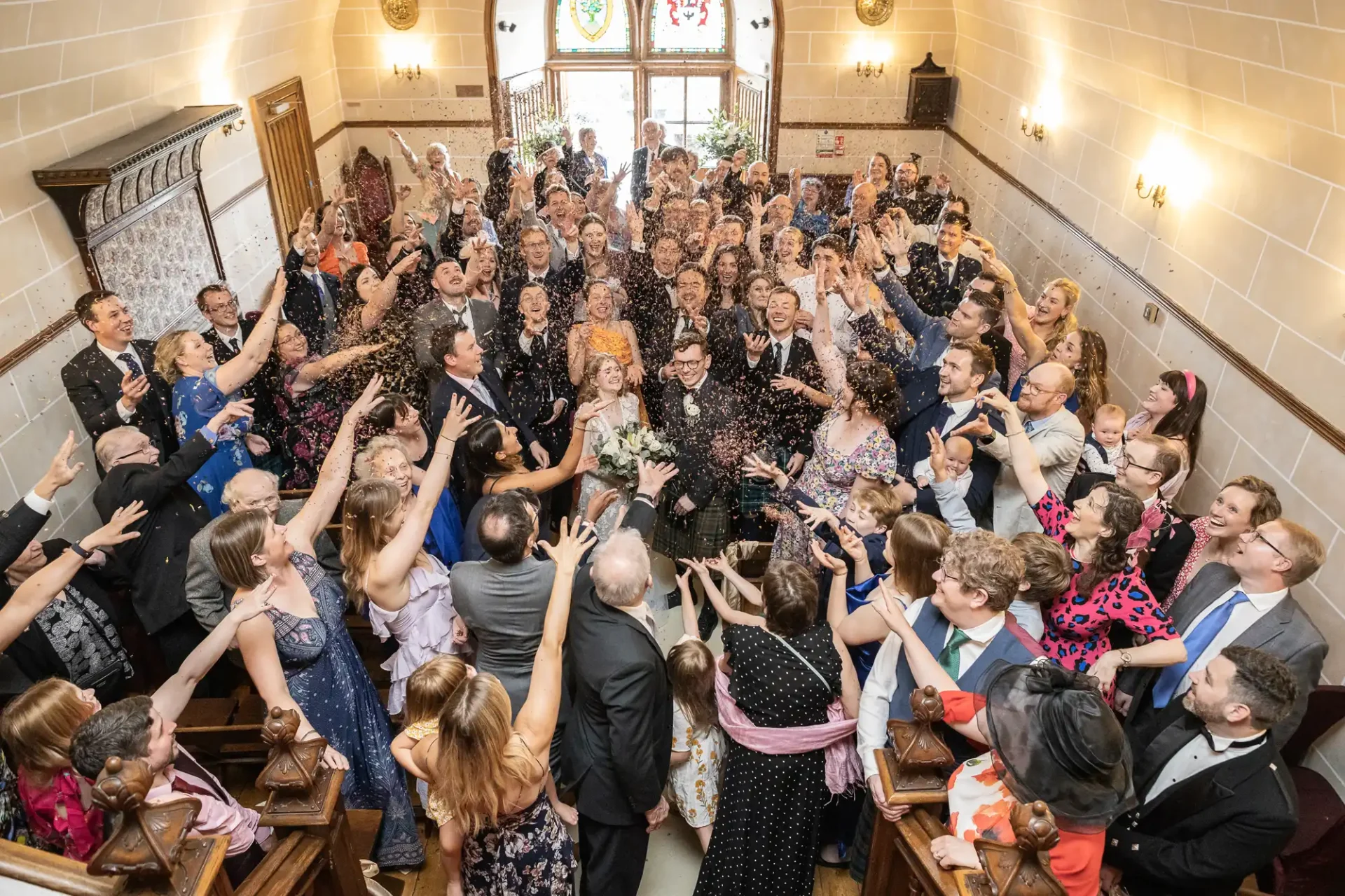 A large group of guests cheer and throw confetti at a newly married couple at an indoor wedding venue. The couple stands in the center, both smiling broadly.