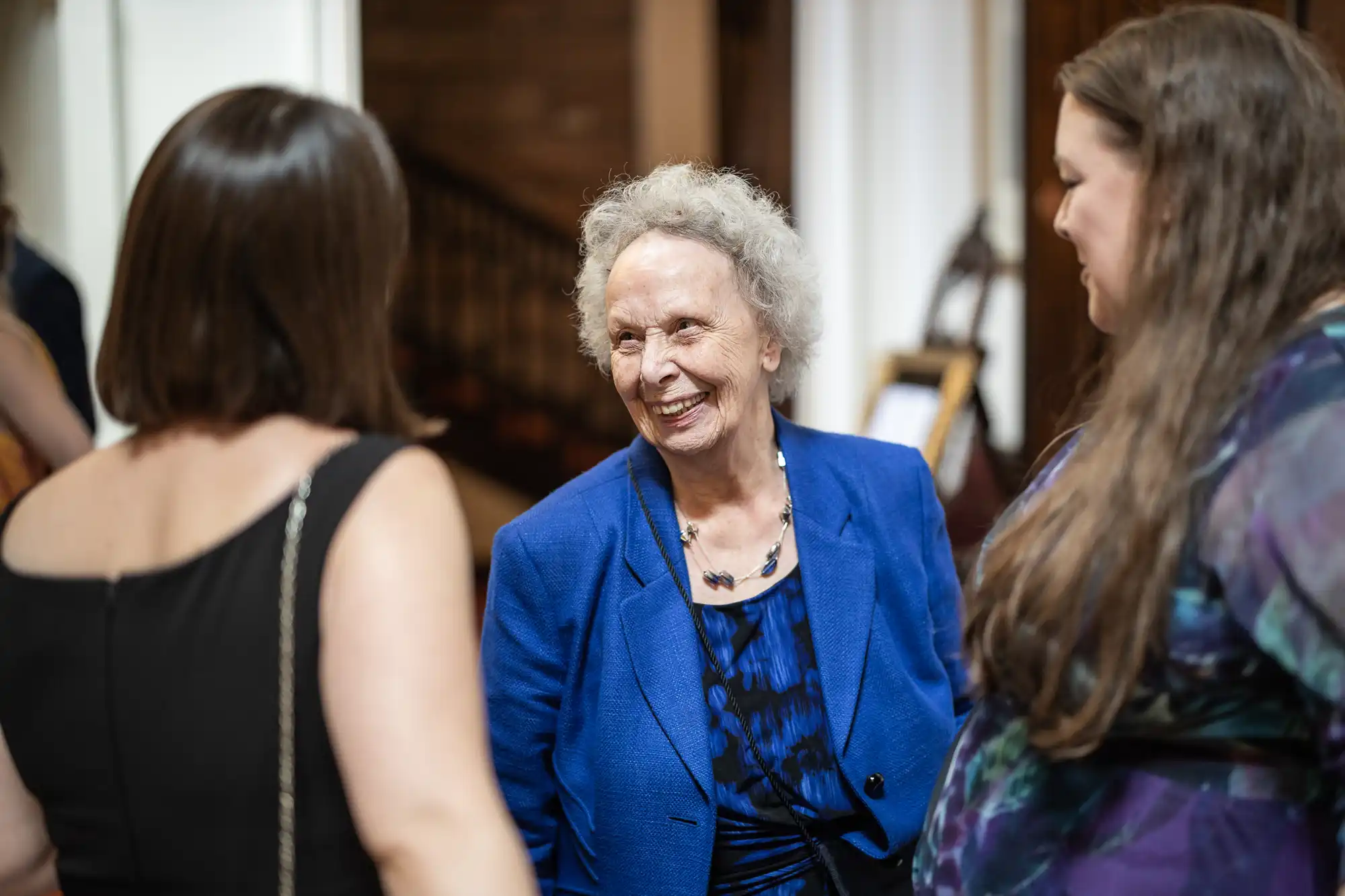 An elderly woman with curly gray hair wearing a blue blazer is talking and smiling with two women at an indoor event.