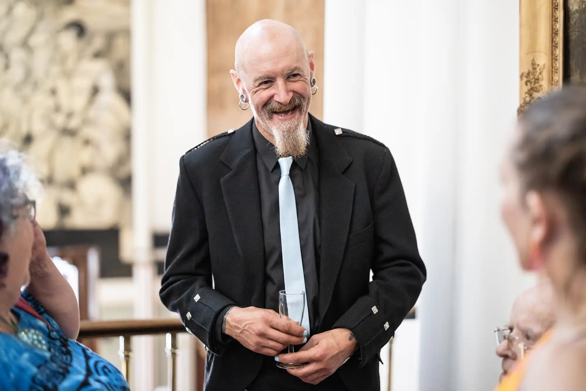 A bald man with a beard and earrings, dressed in a black suit and light blue tie, smiles while holding a glass. He seems to be engaged in conversation with others nearby in a formal setting.