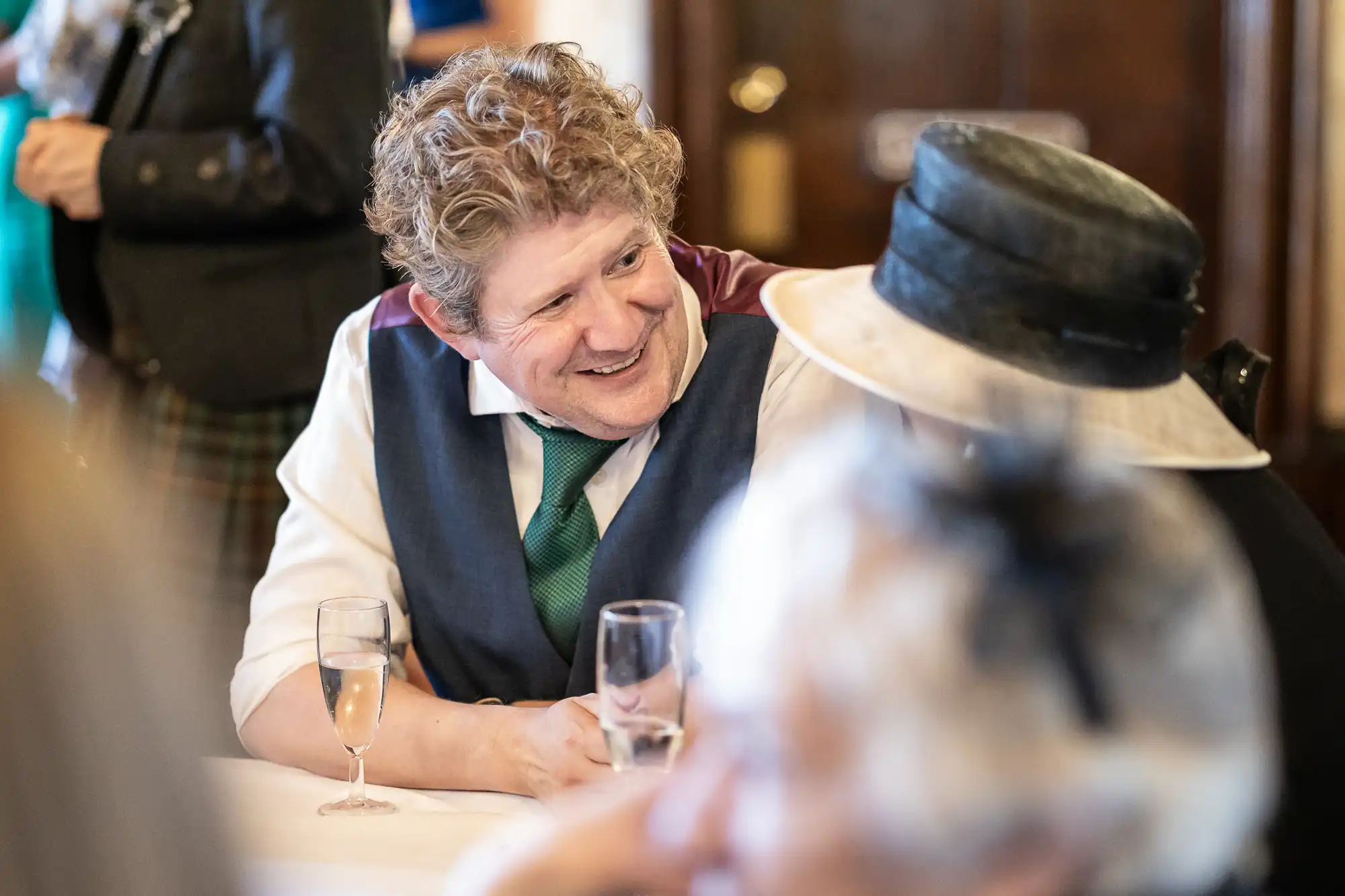 A man with curly hair in formal attire smiles while sitting at a table and holding a glass of champagne, conversing with a person wearing a hat. Other people are seen in the background.