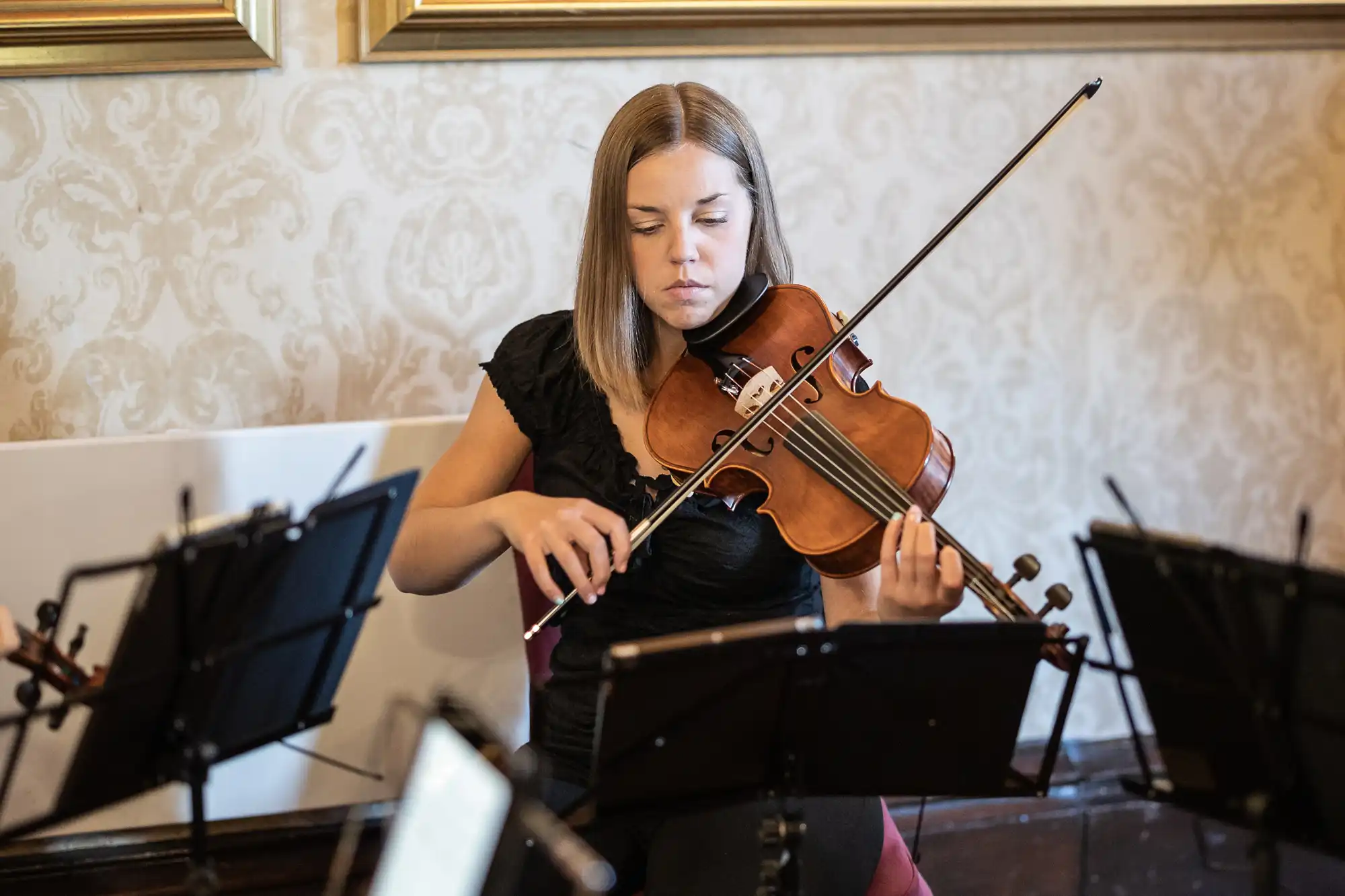 A woman seated and playing a violin, focused on her music sheet, surrounded by music stands in a room with ornate wallpaper and framed pictures.