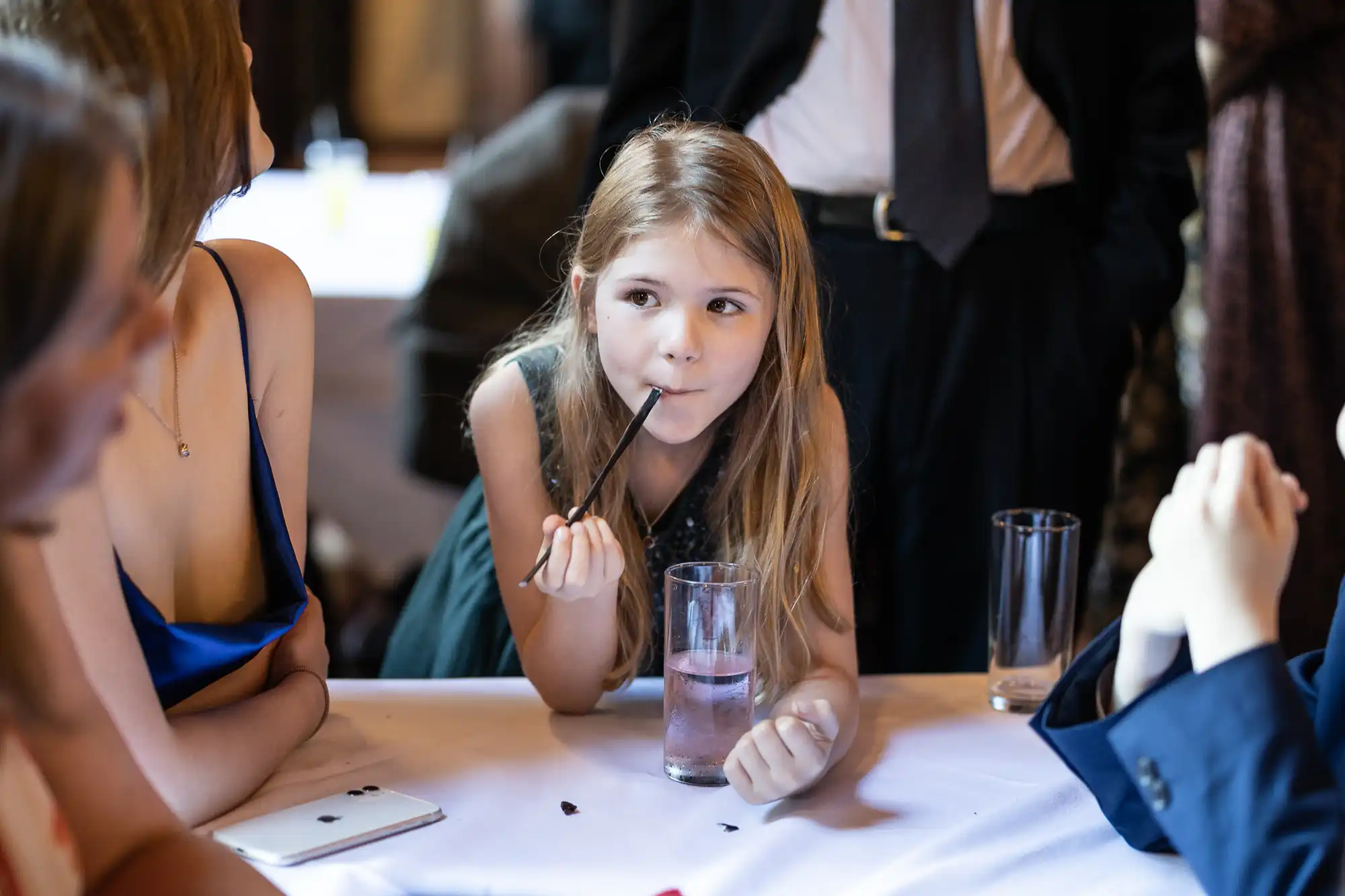 A girl sipping a drink through a straw at a table during a social gathering, with adults around her.