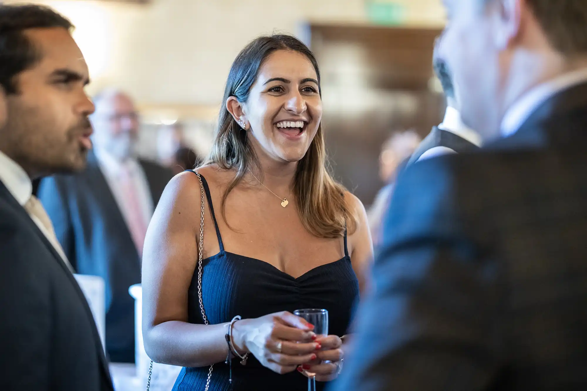 A woman in a black dress is smiling and holding a glass while conversing with others at an indoor social event.