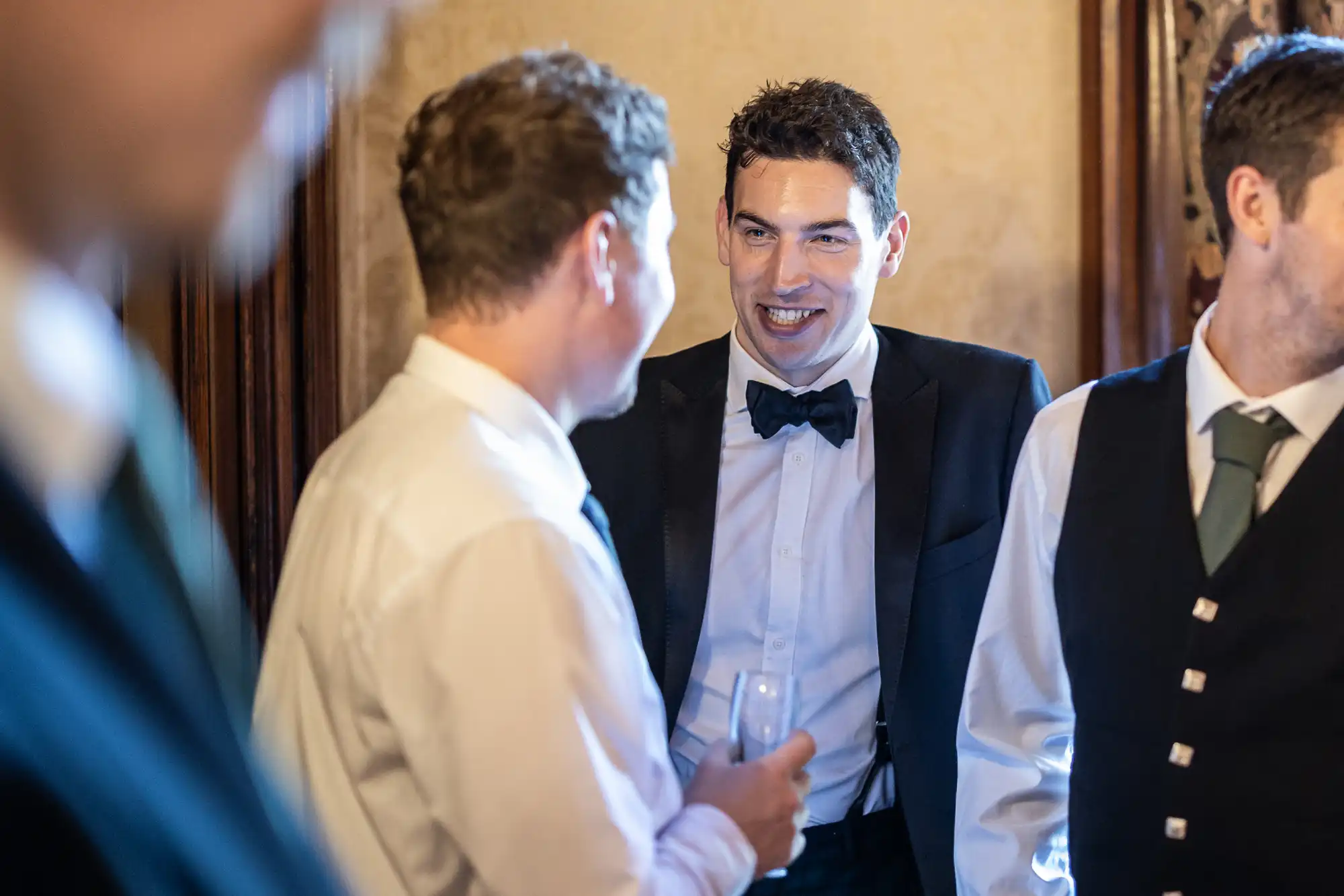 A man in a black tuxedo holds a drink and engages in conversation with two men in white shirts and ties at an indoor gathering.