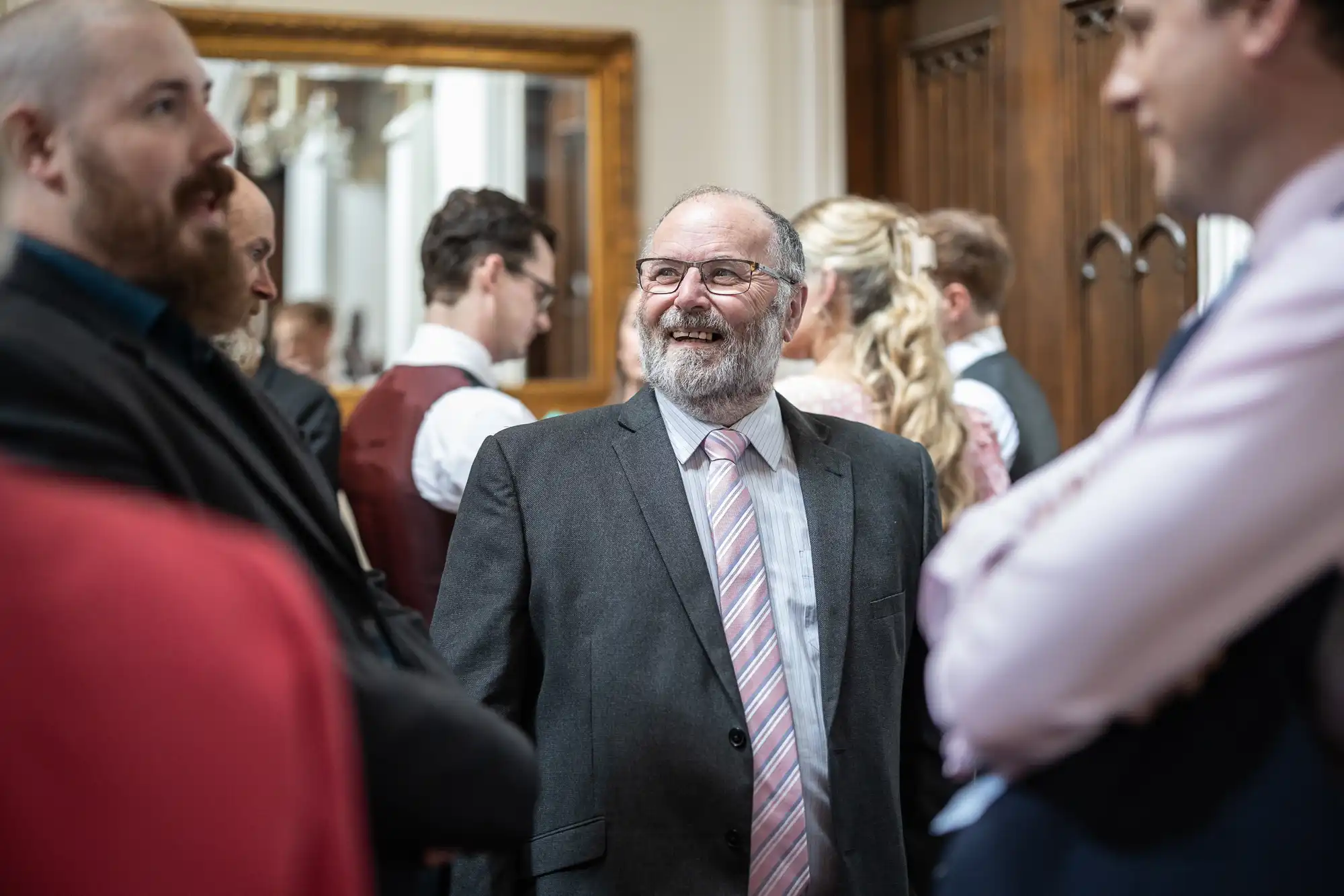 A man in a gray suit and striped tie smiles while talking with others at a formal gathering.