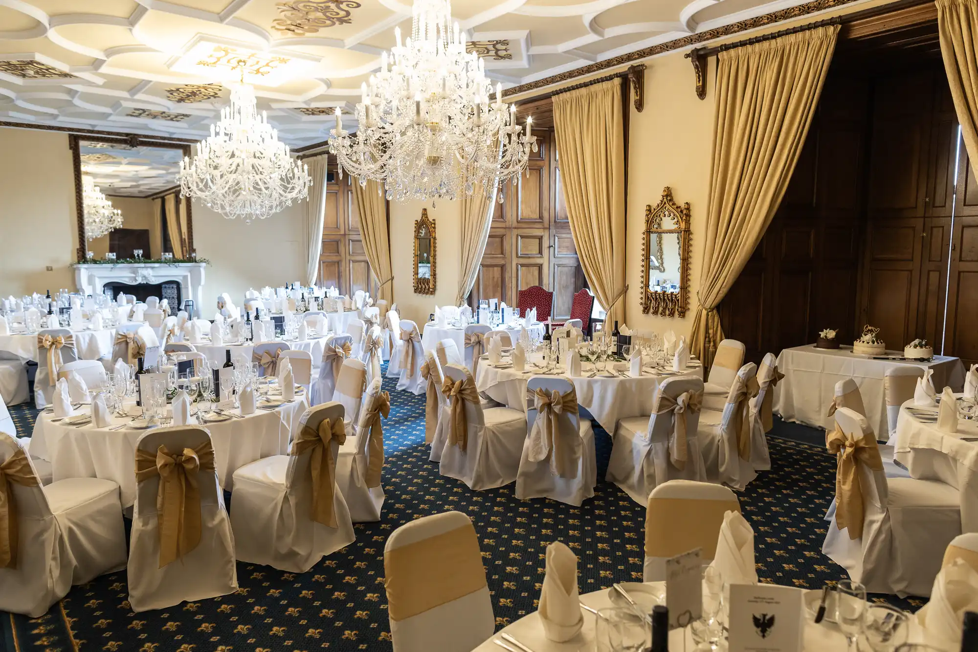 A banquet hall with round tables set for an event, featuring white tablecloths, gold chair sashes, and ornate chandeliers. The room has cream-colored walls, wooden paneling, and large mirrors.