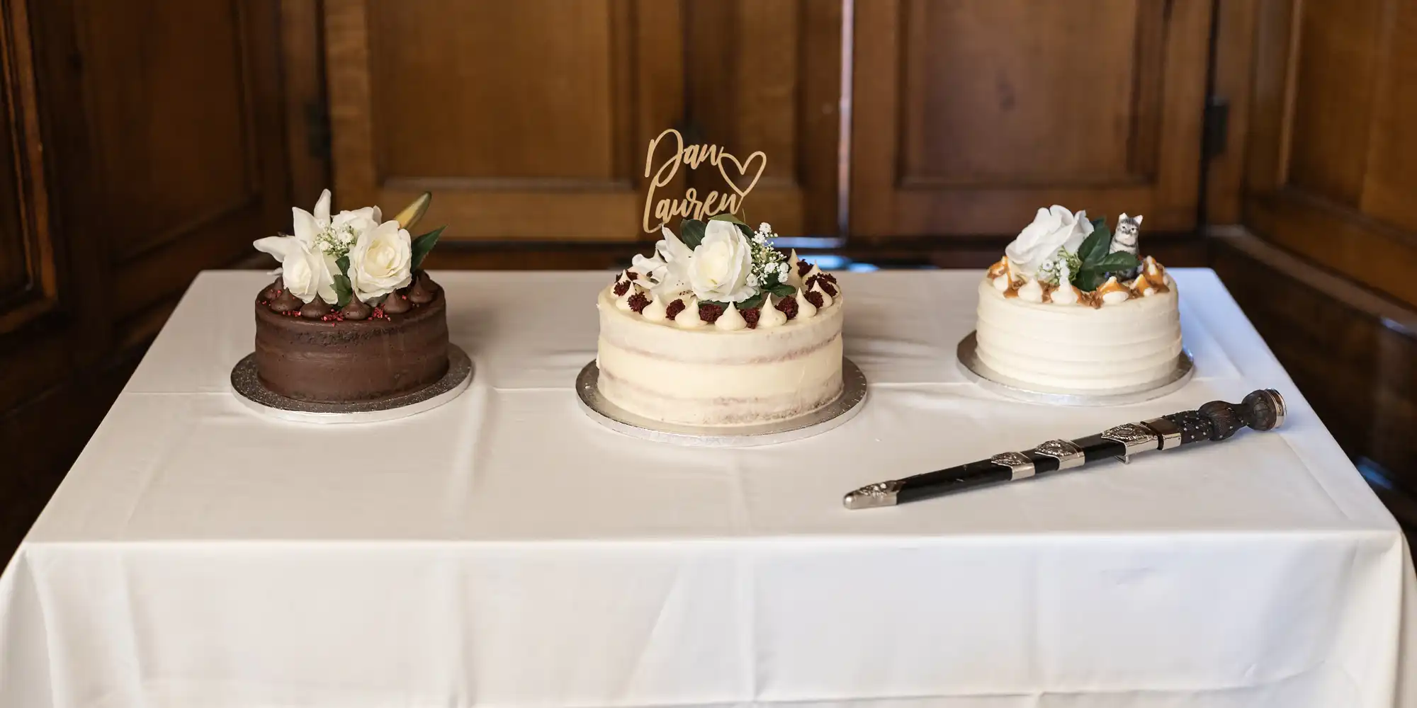 Three decorated cakes sit on a white tablecloth; one chocolate, one with "Pam Lauren" topper, and one with caramel drizzle, alongside a ceremonial sword.