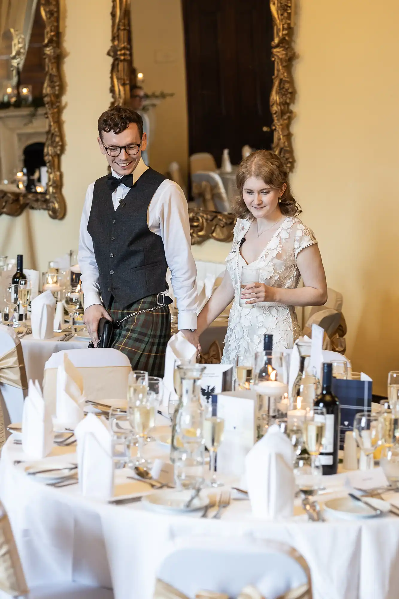 A couple in formal attire, with the man wearing a vest and kilt and the woman in a white dress, walks hand in hand by a dining table set for an event.