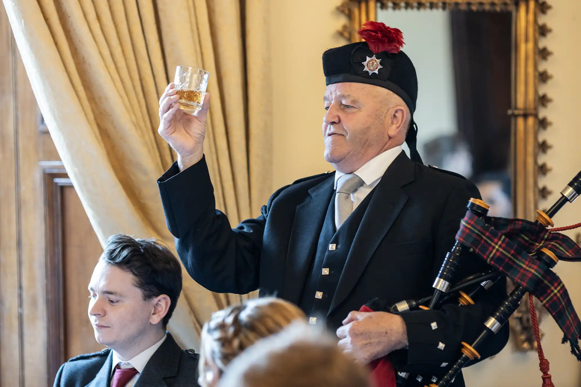 A man in traditional Scottish attire holds up a glass of whisky. He is holding bagpipes in his other arm, and there are people seated around him.