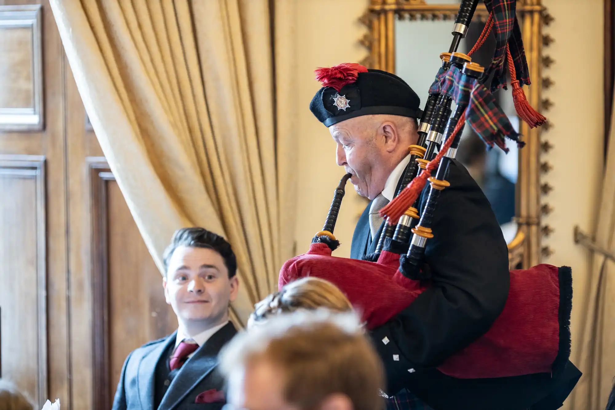 A person plays the bagpipes while wearing traditional Scottish attire in a room with people and a large mirror in the background. A man in a suit looks on.