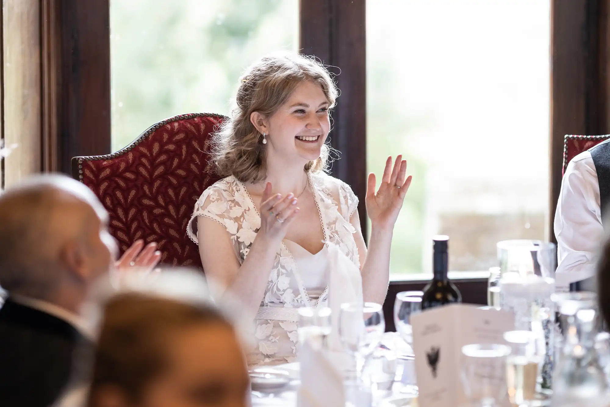 A smiling woman with light hair claps while sitting at a table set with glasses and bottles during an event.