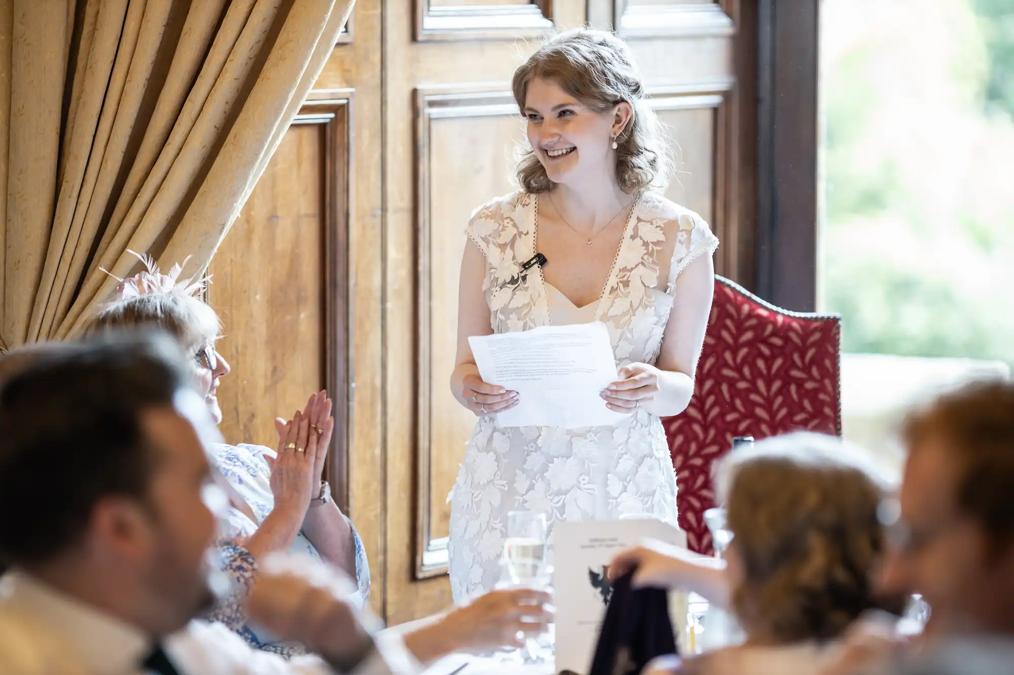 A woman in a white dress stands holding a piece of paper, speaking to a seated group of people in an elegant room. One seated woman claps while others listen attentively.