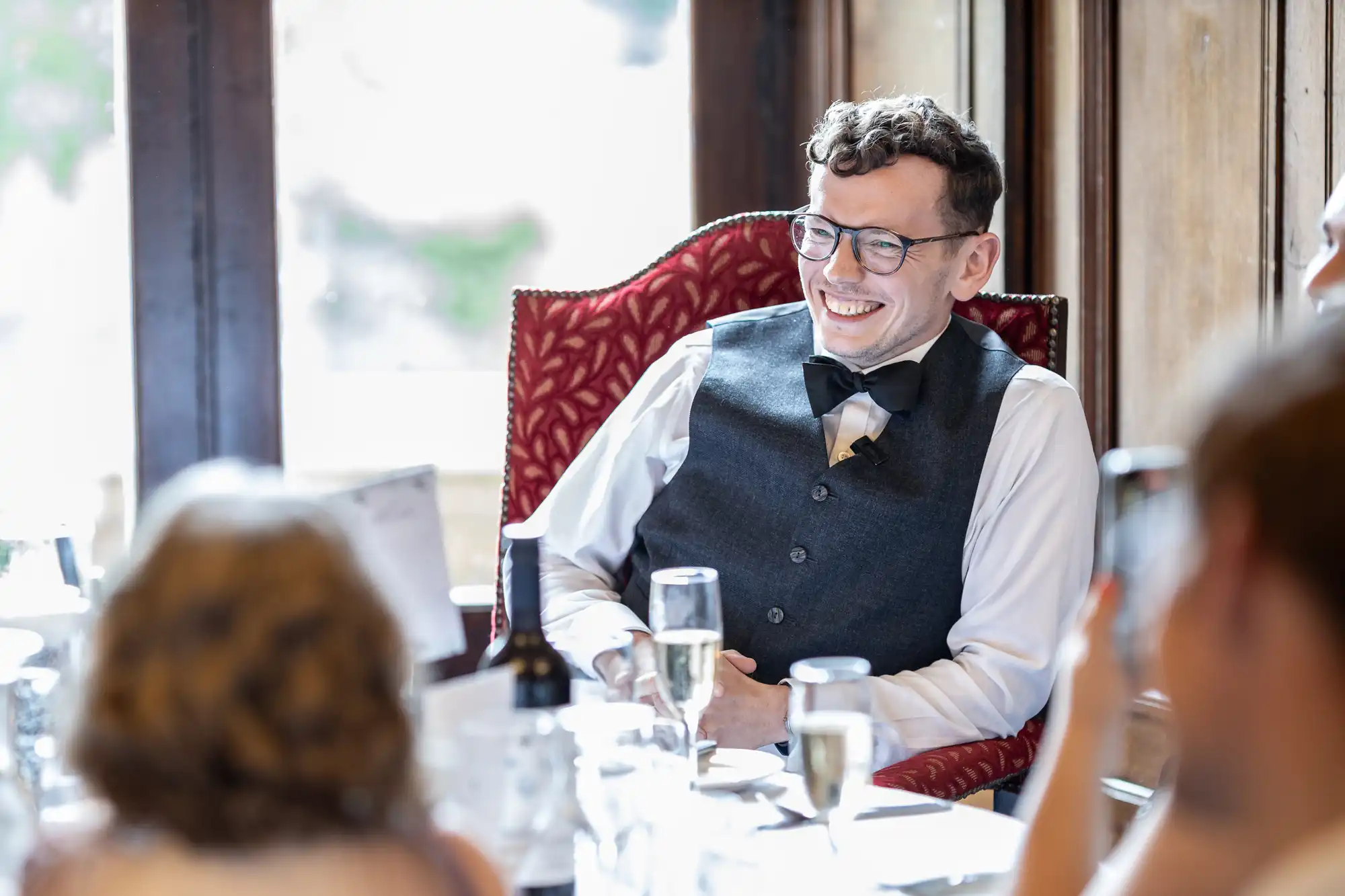 A man wearing glasses, a bow tie, and a vest is sitting and smiling at a table during a social event, with blurred people and a window in the background.