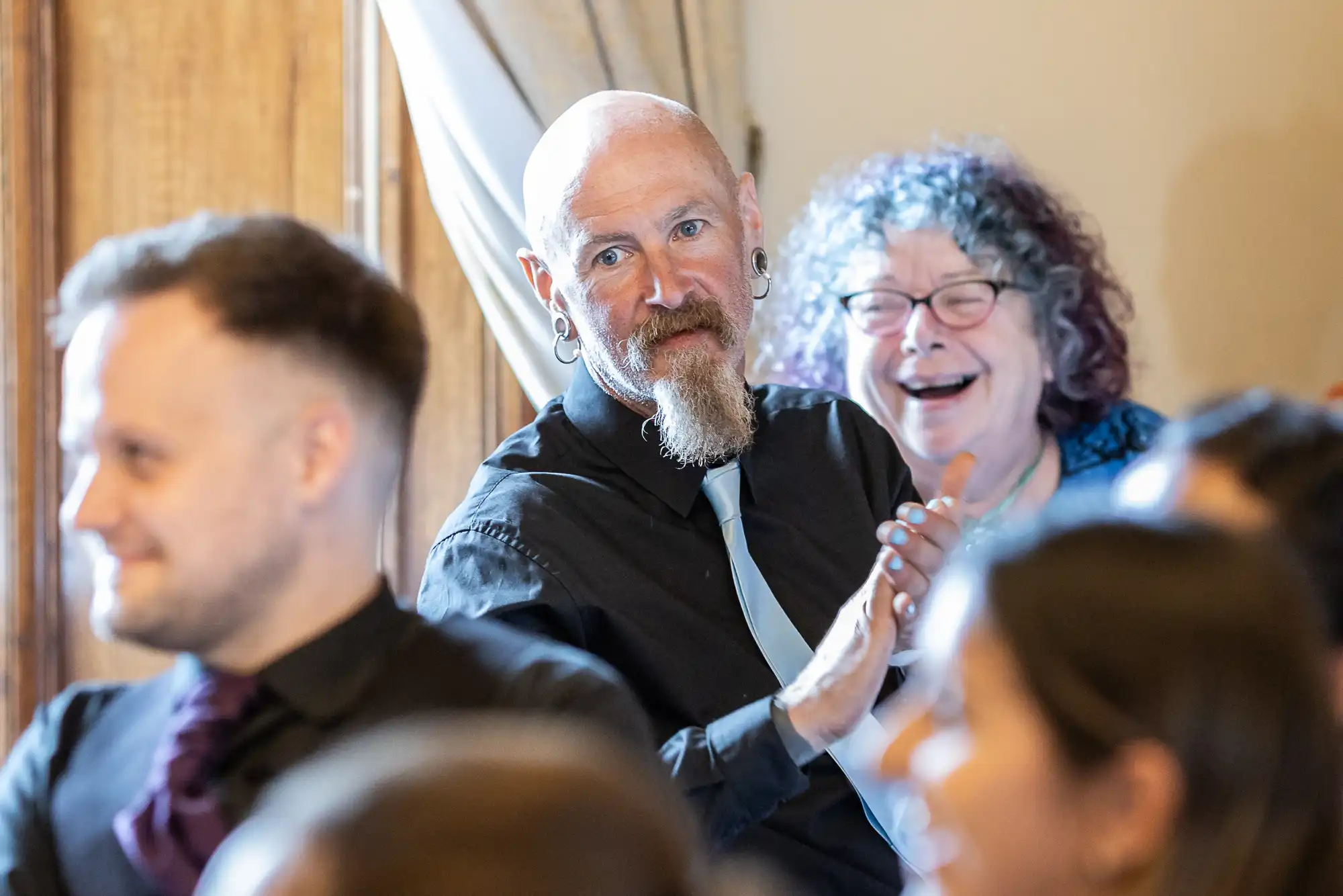 A man with a beard and earrings, and a woman with curly hair and glasses, smile and clap among seated attendees in a room.