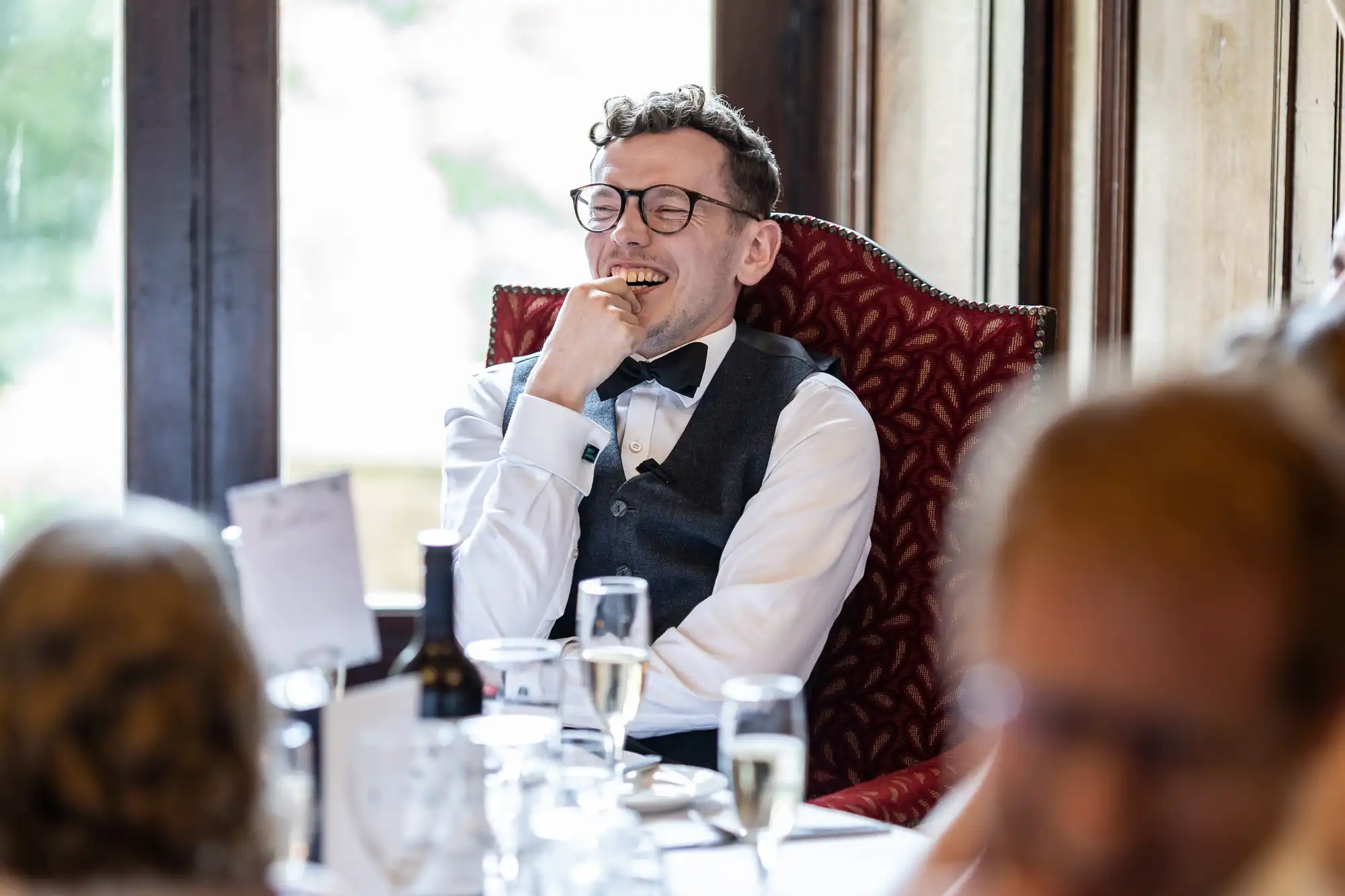 A man wearing glasses and a formal vest with a bow tie is sitting on a red chair, laughing while holding a cracker, at a dining table set with glasses and a bottle.