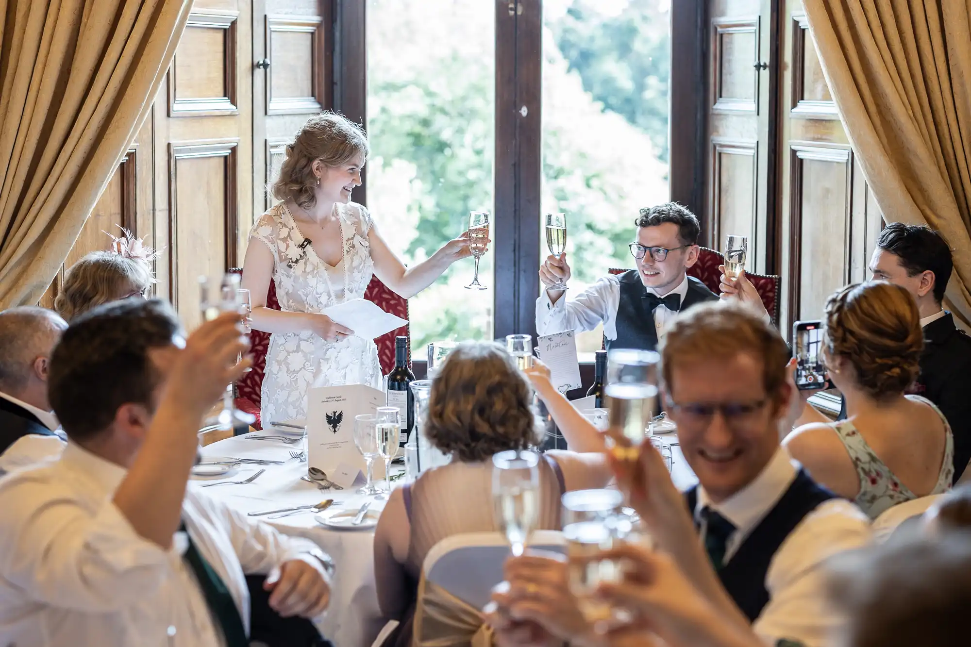 A bride and groom raise their glasses in a toast while surrounded by guests seated at a round table in a well-lit room with large windows and curtains.