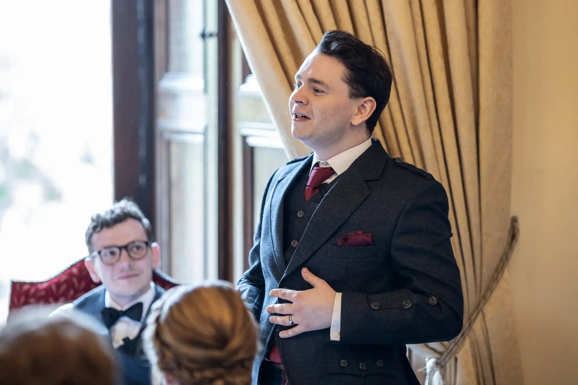 A man in a dark suit and red tie is speaking in front of a group of people in a room with curtains and a large window. Another man with glasses is seated and listening attentively.