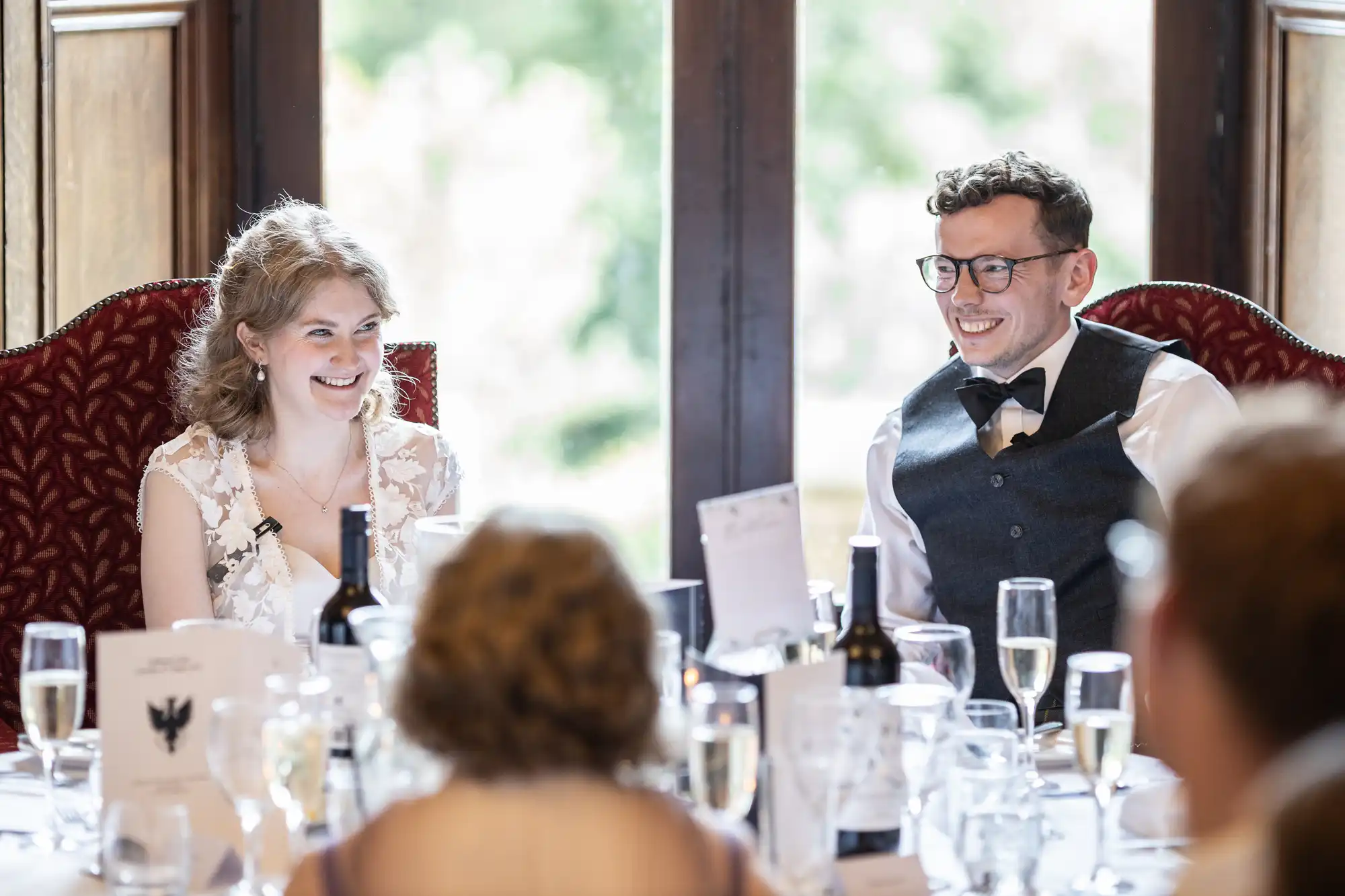 A bride and groom sit and smile at a table adorned with wine glasses and decor, during a celebration.