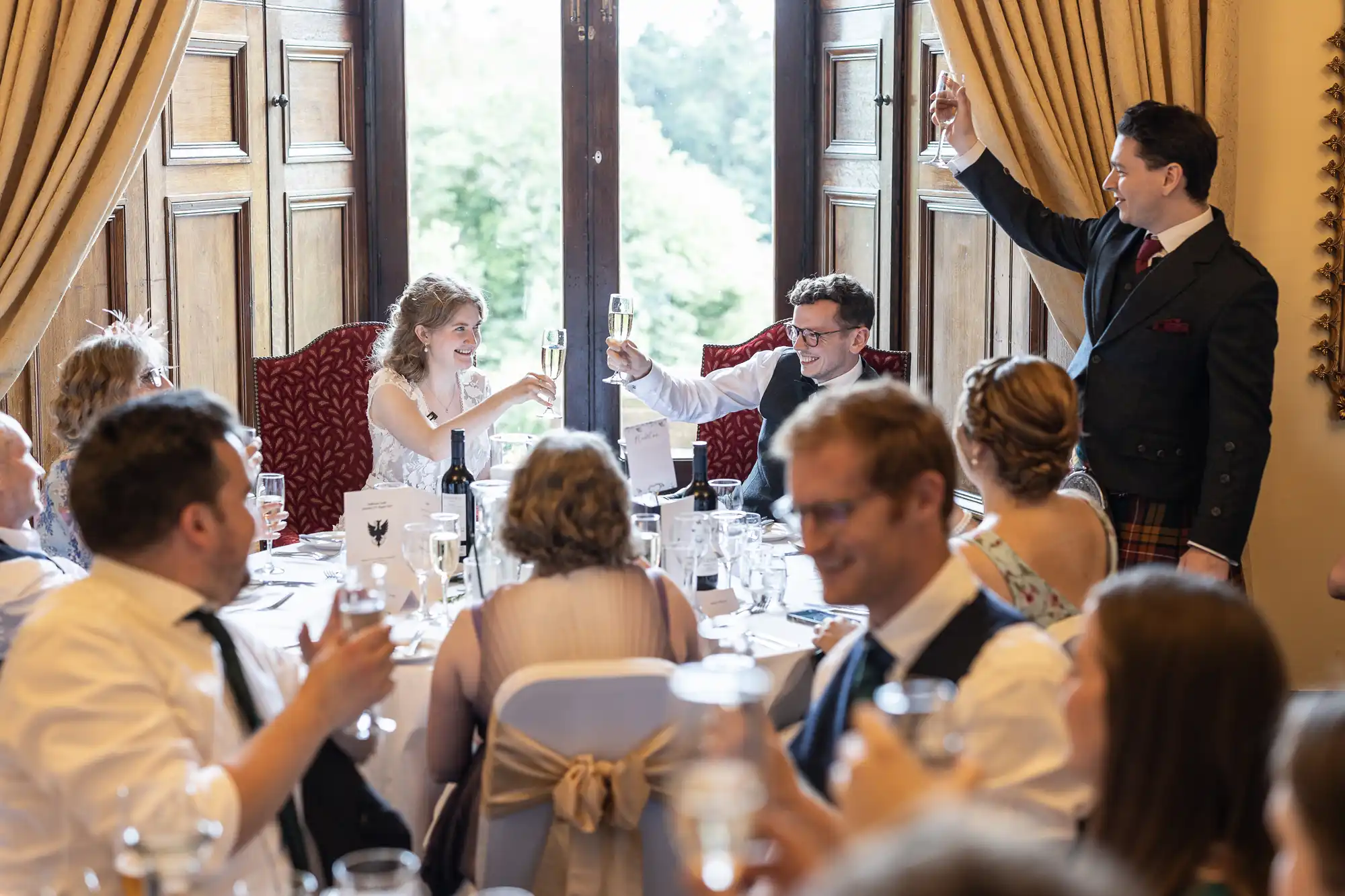 A group of people seated around a dining table raising their glasses in a toast, with a couple seated at the head of the table. The setting appears to be a formal gathering or celebration.