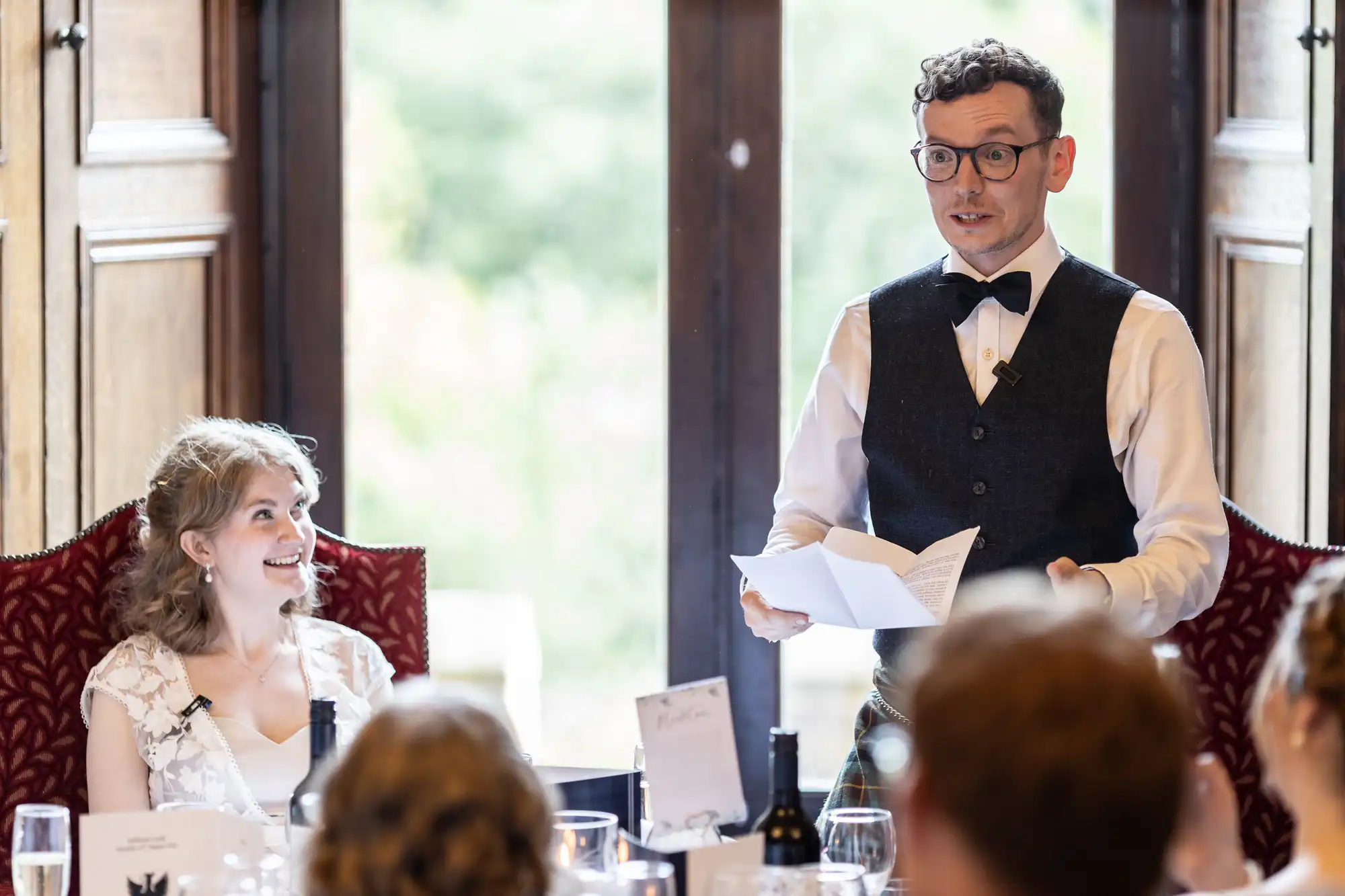 A man in a bow tie and vest speaks while holding papers, with a woman seated next to him, smiling, at a formal gathering.