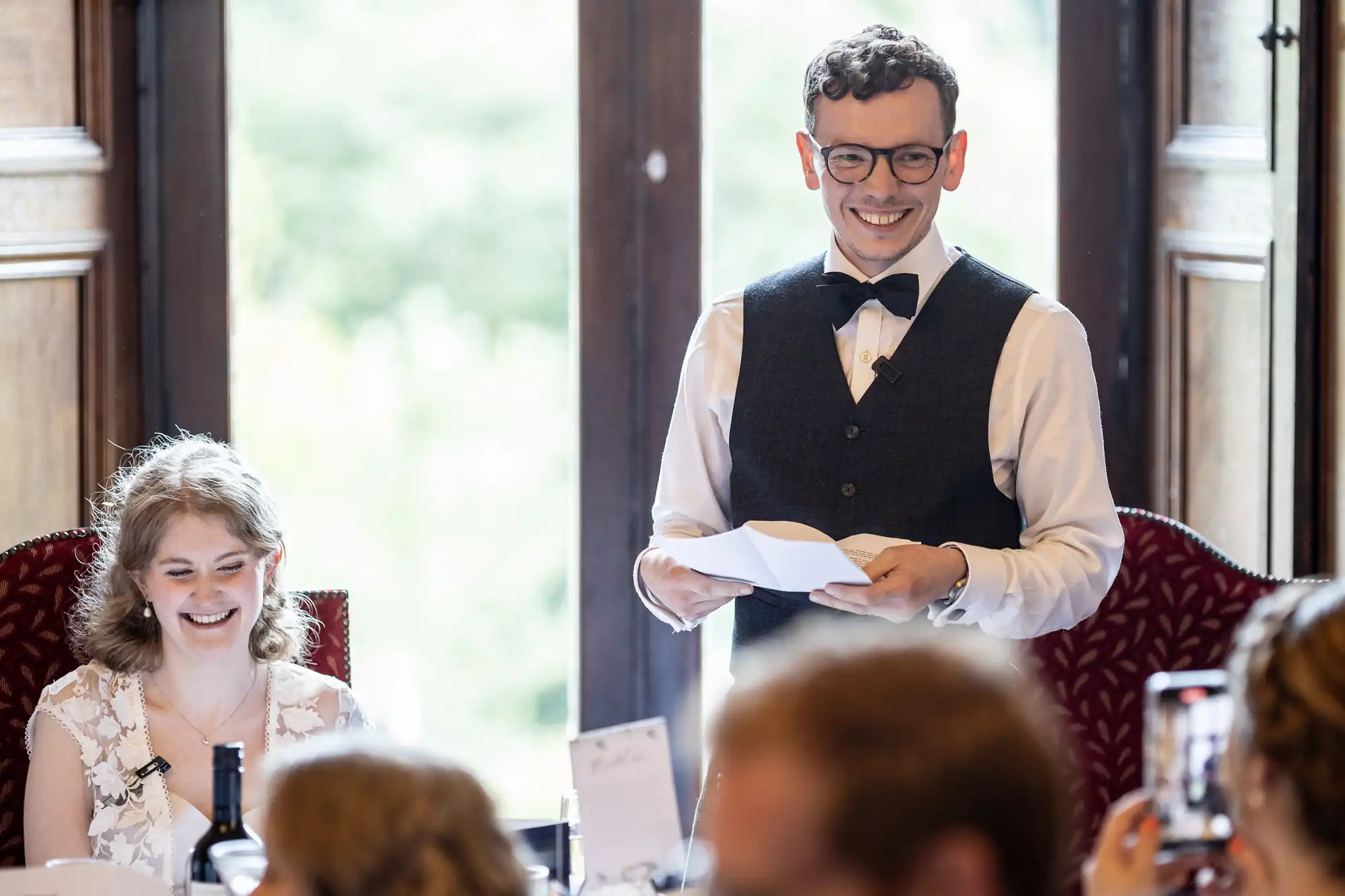 A man in a bow tie and vest speaks while holding papers. A woman with curly hair sits smiling next to him. Both are inside a room with wooden paneling and large windows in the background.