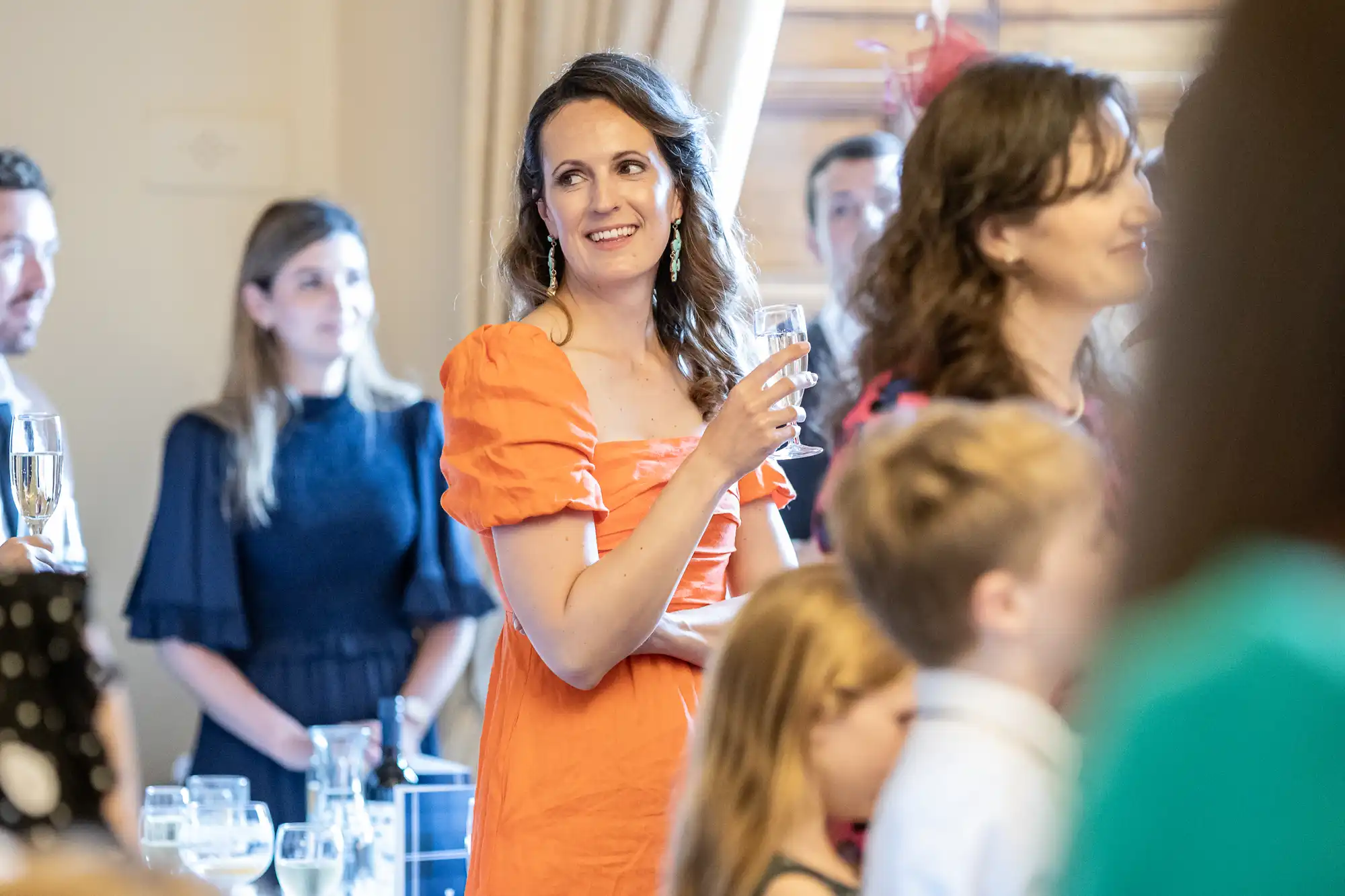 A woman in an orange dress holds a glass and smiles in a room with other people. Some are standing while others are sitting, engaging in conversations.