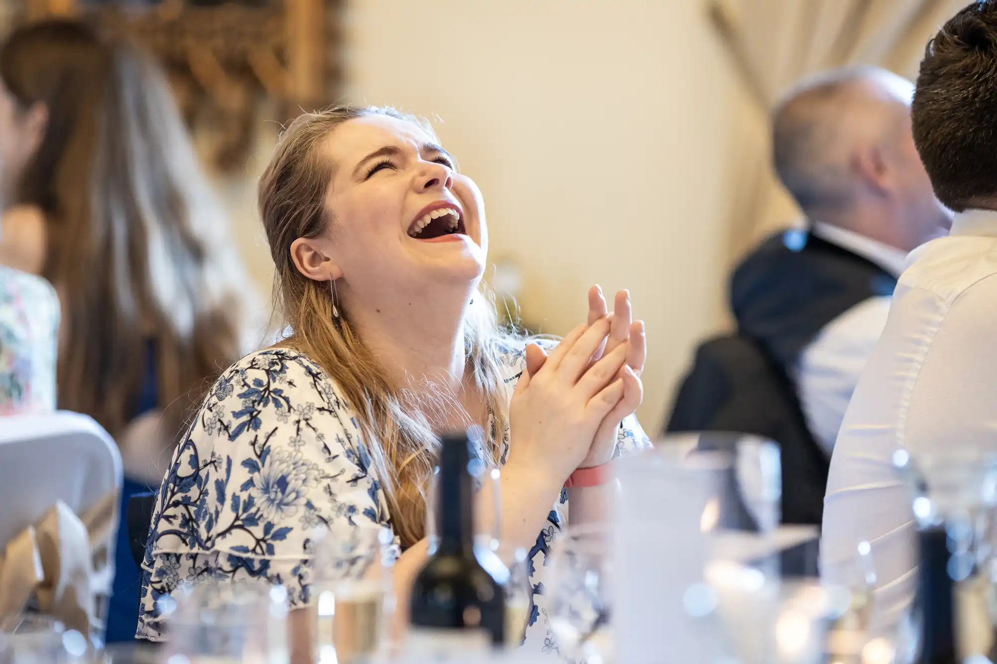 A woman in a floral dress, seated at a table with drinks, laughs with hands clasped. Other people are seated around her.