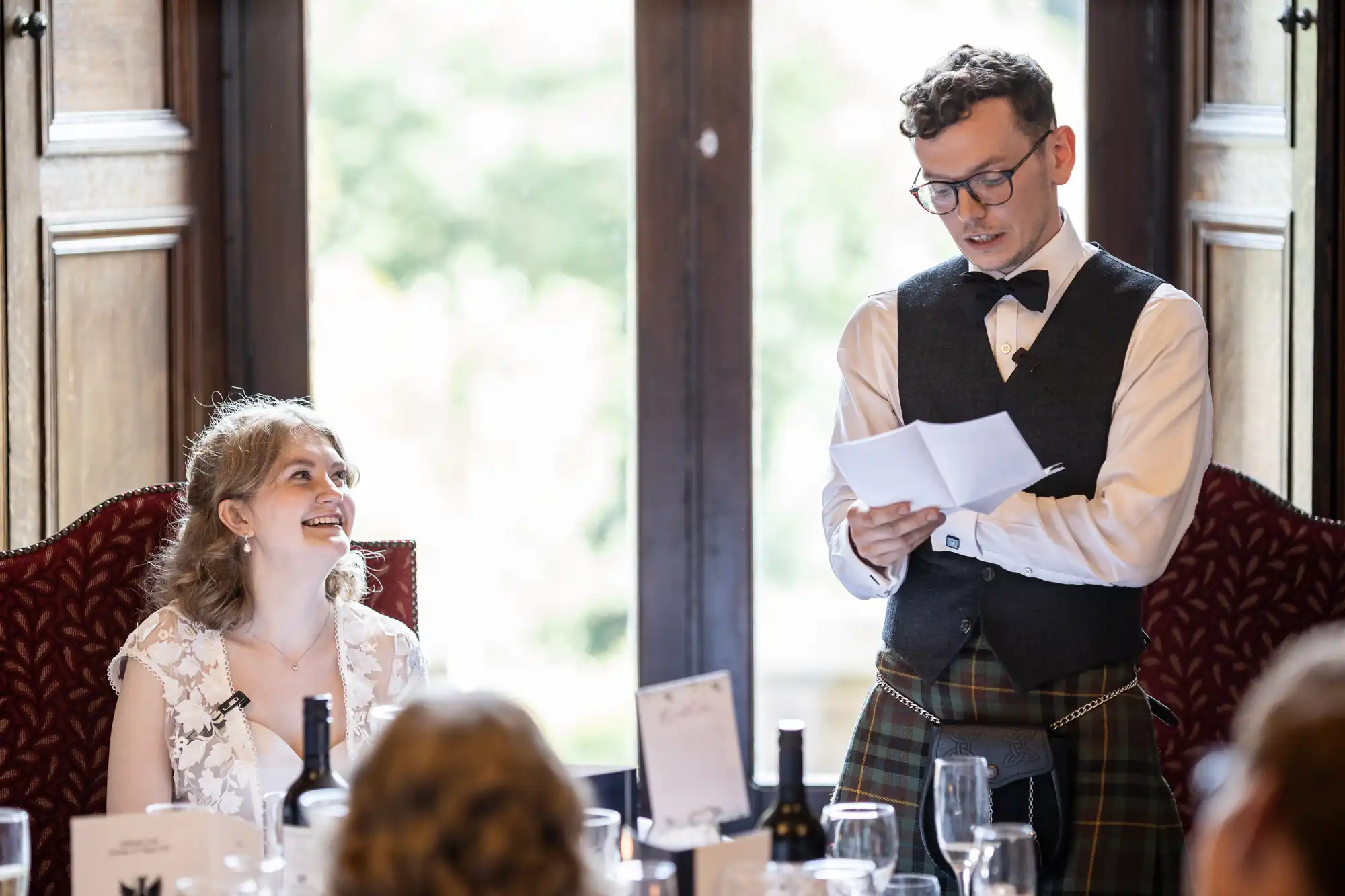 A person smiles while seated as another person, holding a paper and wearing glasses, a bow tie, and a kilt, stands and speaks.