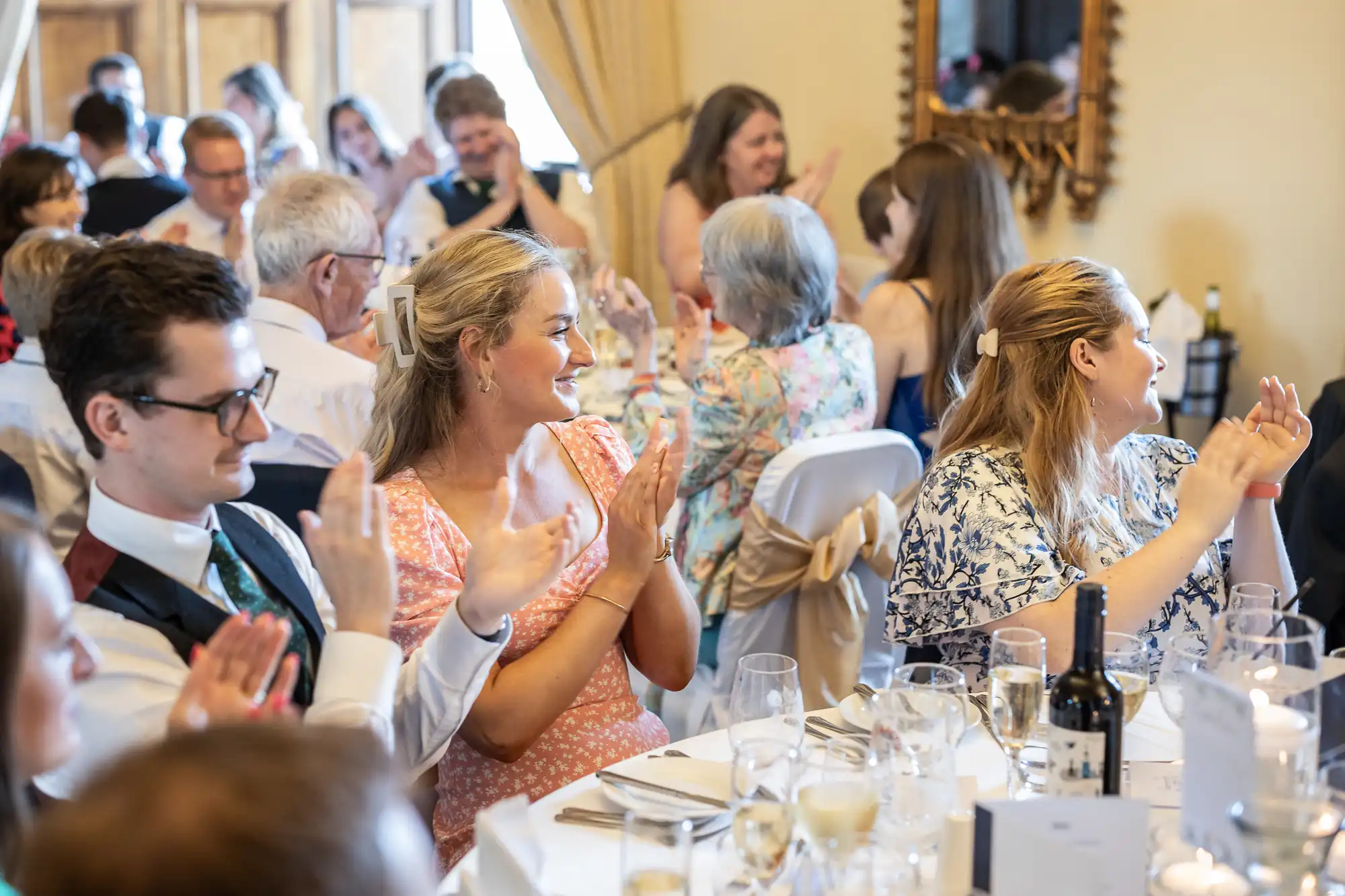 A group of people seated at a formal event, clapping and smiling. The tables are set with white linens, glassware, and bottles of wine. A large mirror and decorative curtains are in the background.